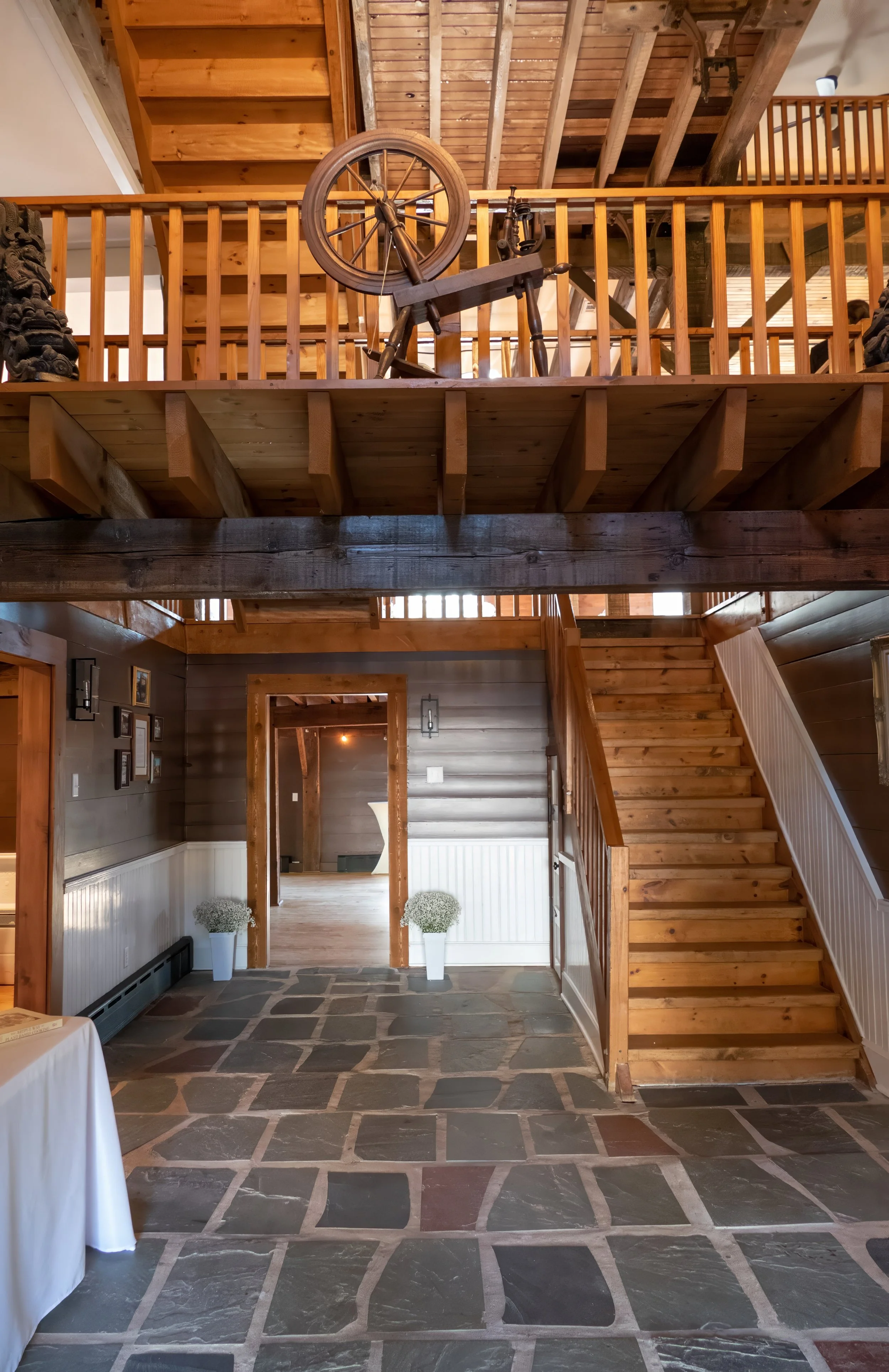 Interior view of the foyer of a rustic wooden event venue with a stone floor, wooden walls, stairs, and dance hall visible through a doorway, with decorative vase and framed pictures.