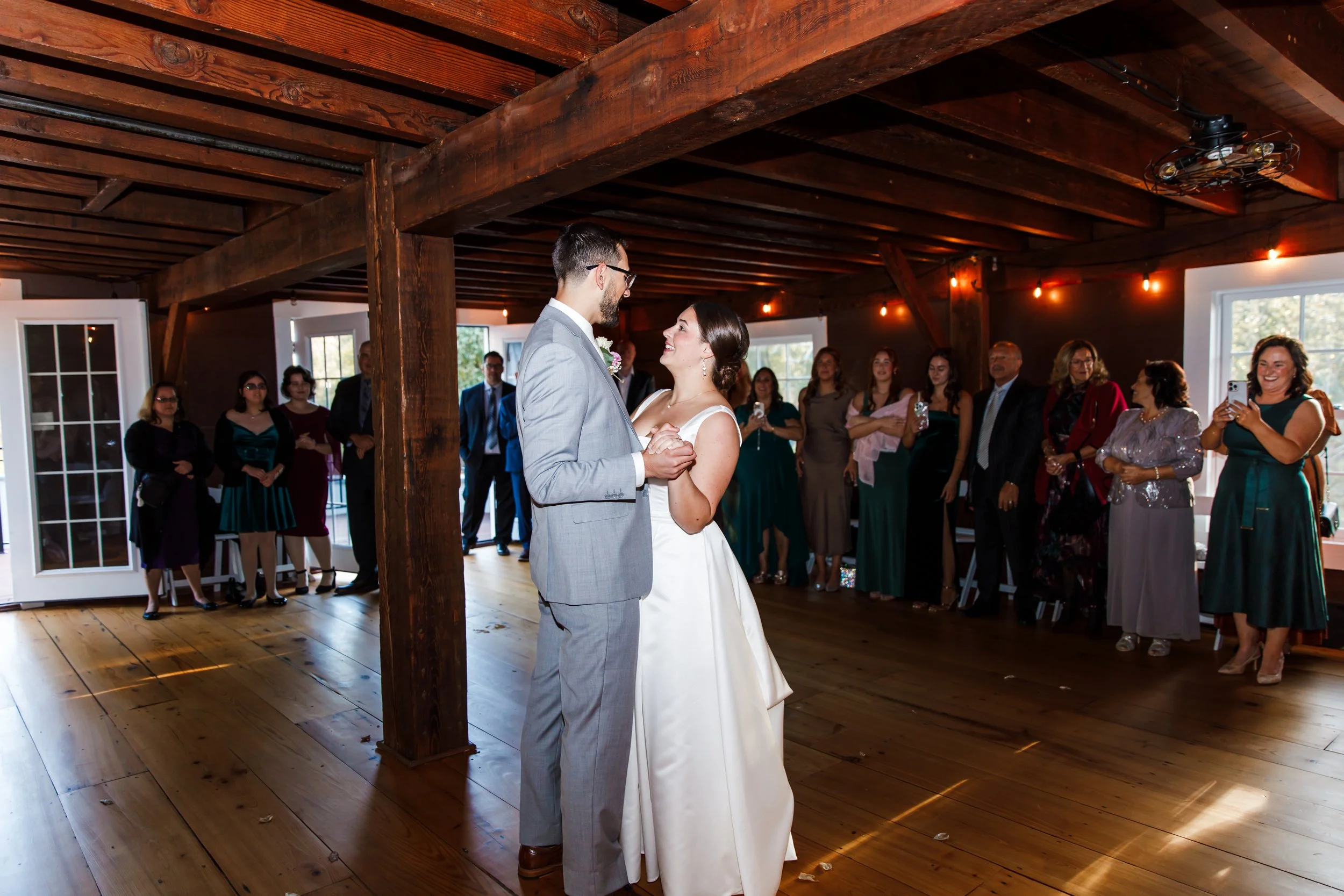 A bride and groom having their first dance at a wedding reception, with family and friends watching and taking photos inside a rustic wooden hall.