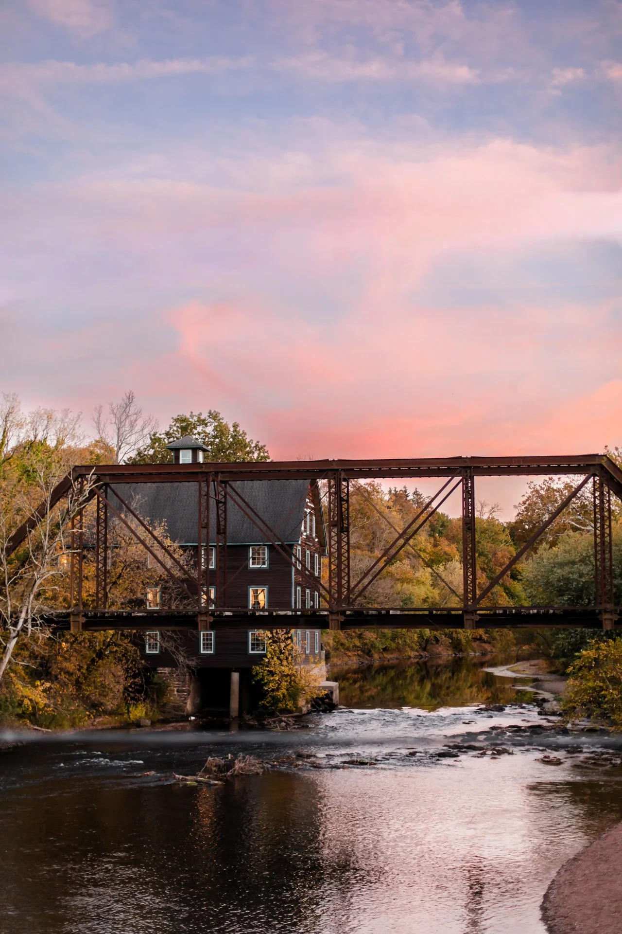 A historic watermill with a dark wooden exterior and multiple windows, situated under a metal bridge spanning a flowing river during sunset with colorful sky and autumn trees surrounding the scene.