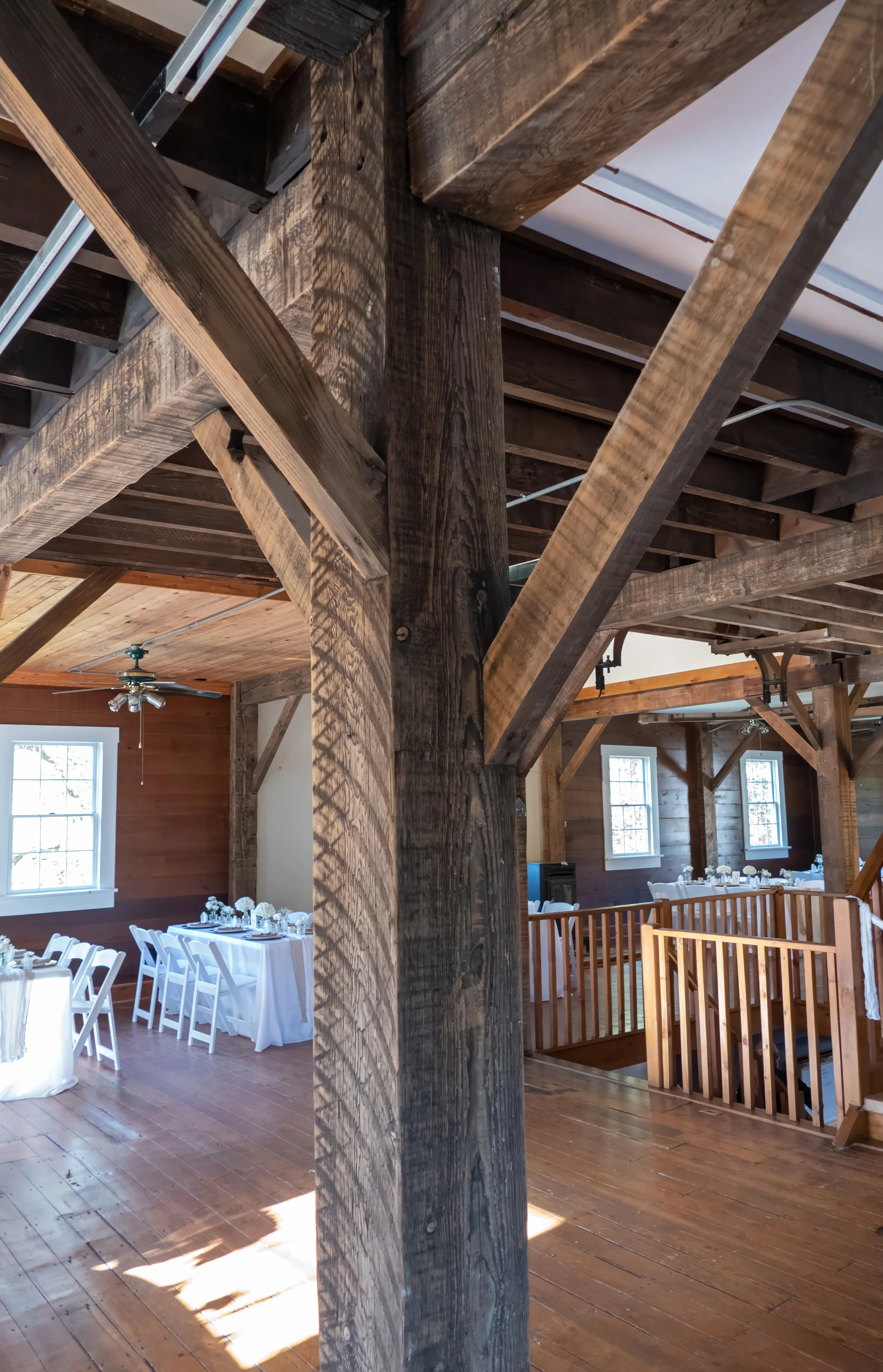 Interior of a rustic venue with wooden beams, white tables with tableware and flowers, wooden floors, and windows.