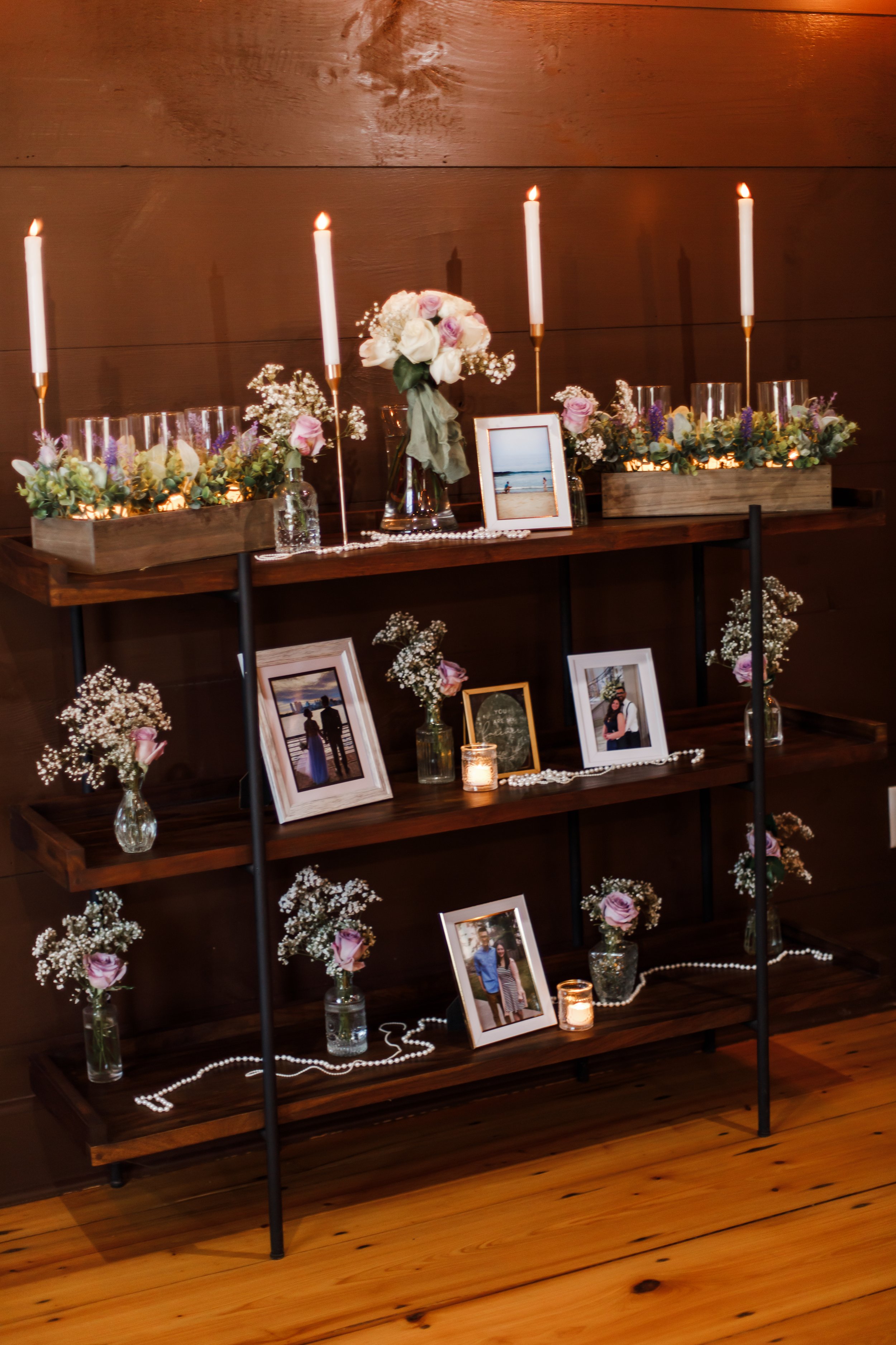 A three-tier wooden shelf memorializing loved ones, decorated with candles, flowers, framed photos, and decorative pearls, against a dark wooden wall.
