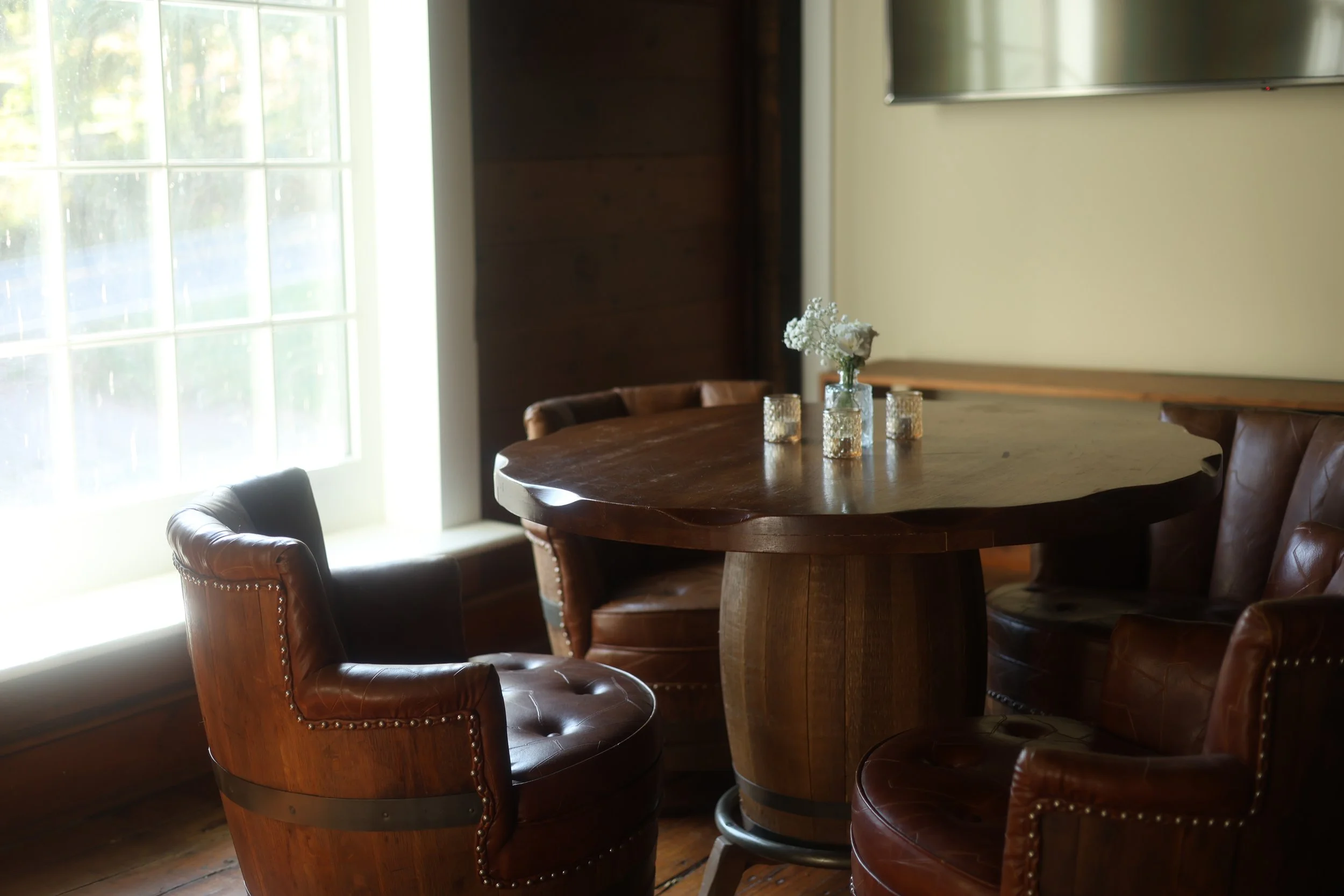 A wooden dining table at an event venue with a floral centerpiece and candles, surrounded by leather chairs, near a window with natural light.