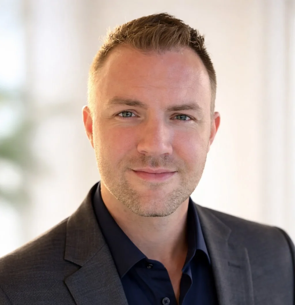 A portrait of a man with short brown hair, light skin, wearing a dark suit jacket and navy shirt, smiling softly, with a blurred indoor background.