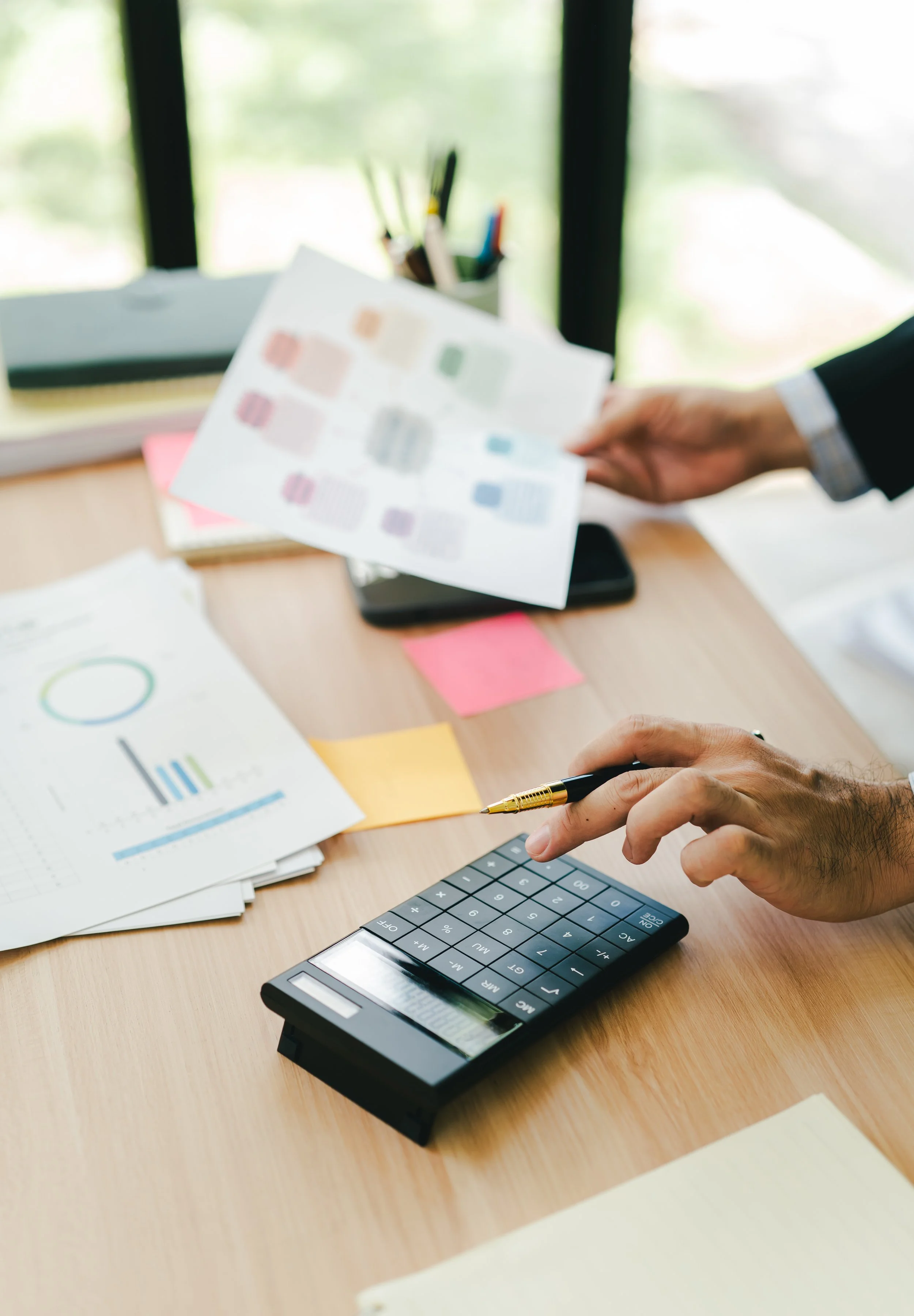 Business person using calculator and holding a color chart at a cluttered wooden desk.