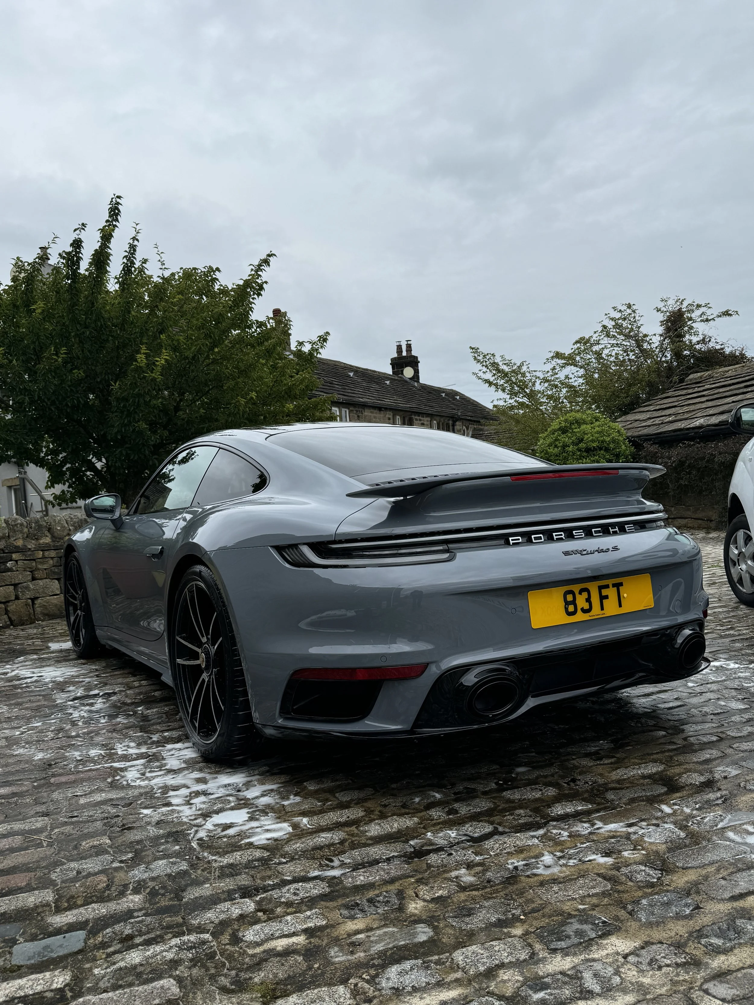 A gray Porsche 911 Turbo S parked on a cobblestone driveway with a yellow license plate reading '83 FT', surrounded by trees and houses on a cloudy day.