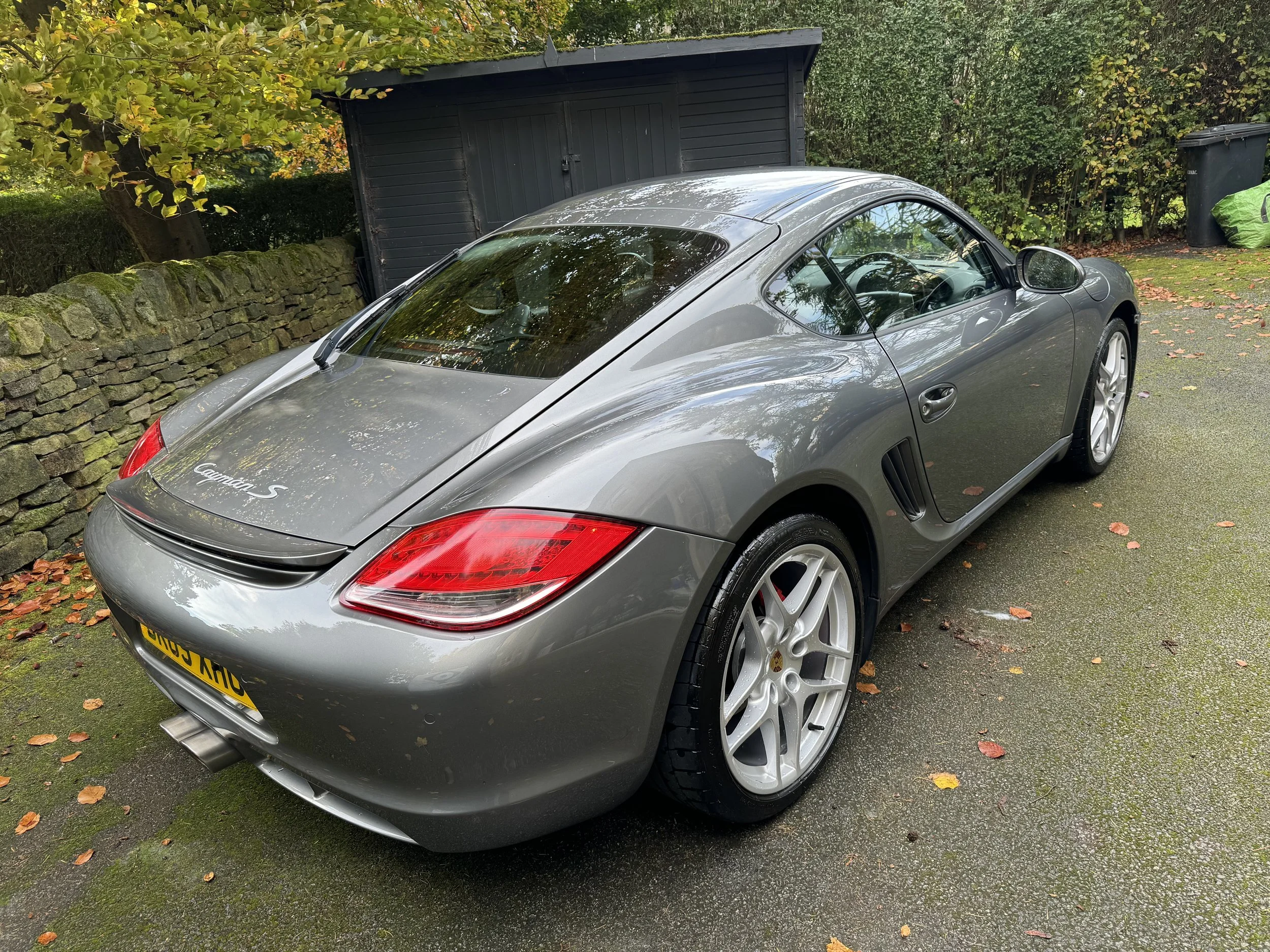 A silver Porsche Cayman S parked on a driveway next to a stone wall and greenery.
