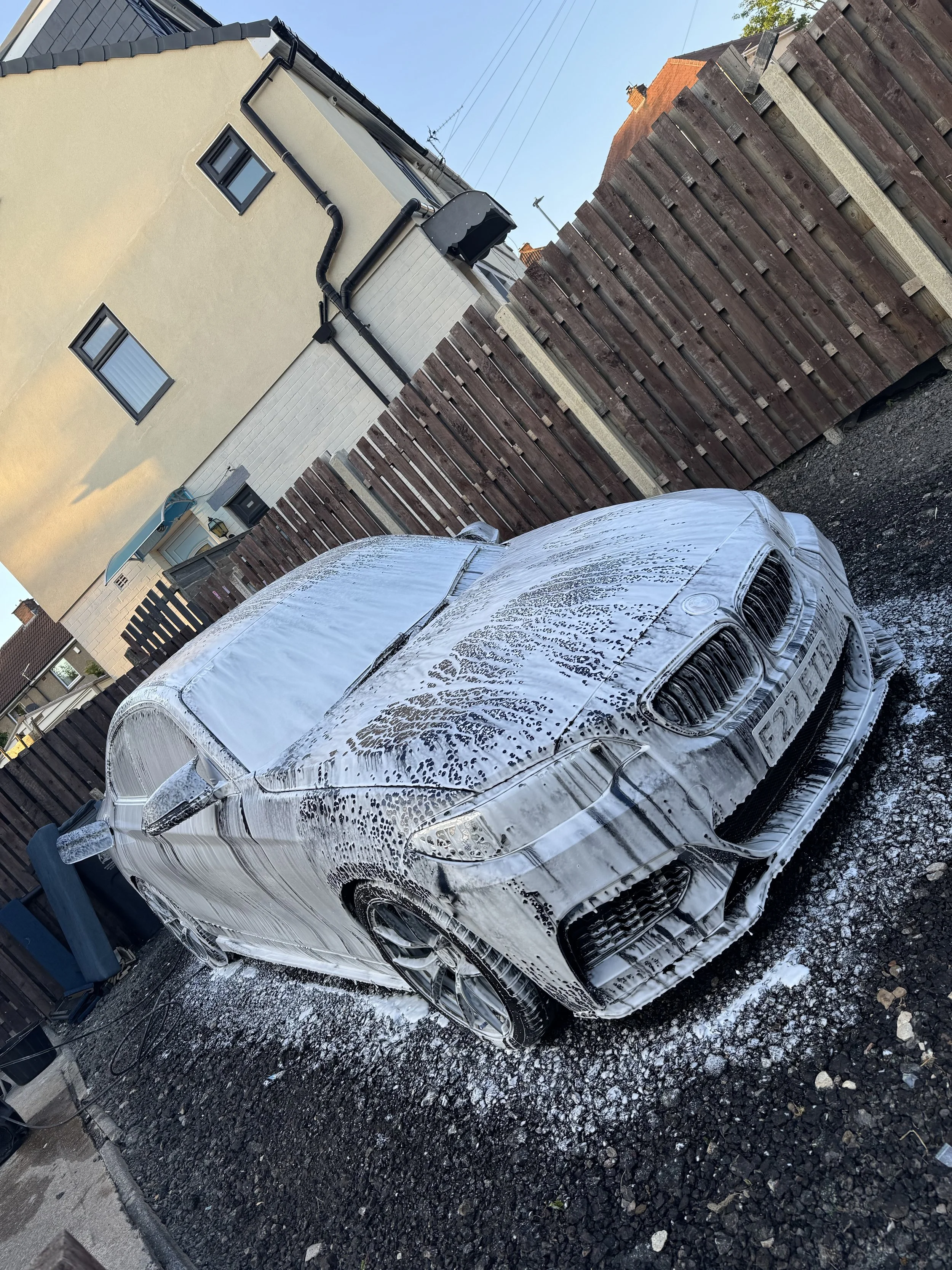 A white car covered in soap suds during a car wash, parked on gravel next to a wooden fence and residential houses.