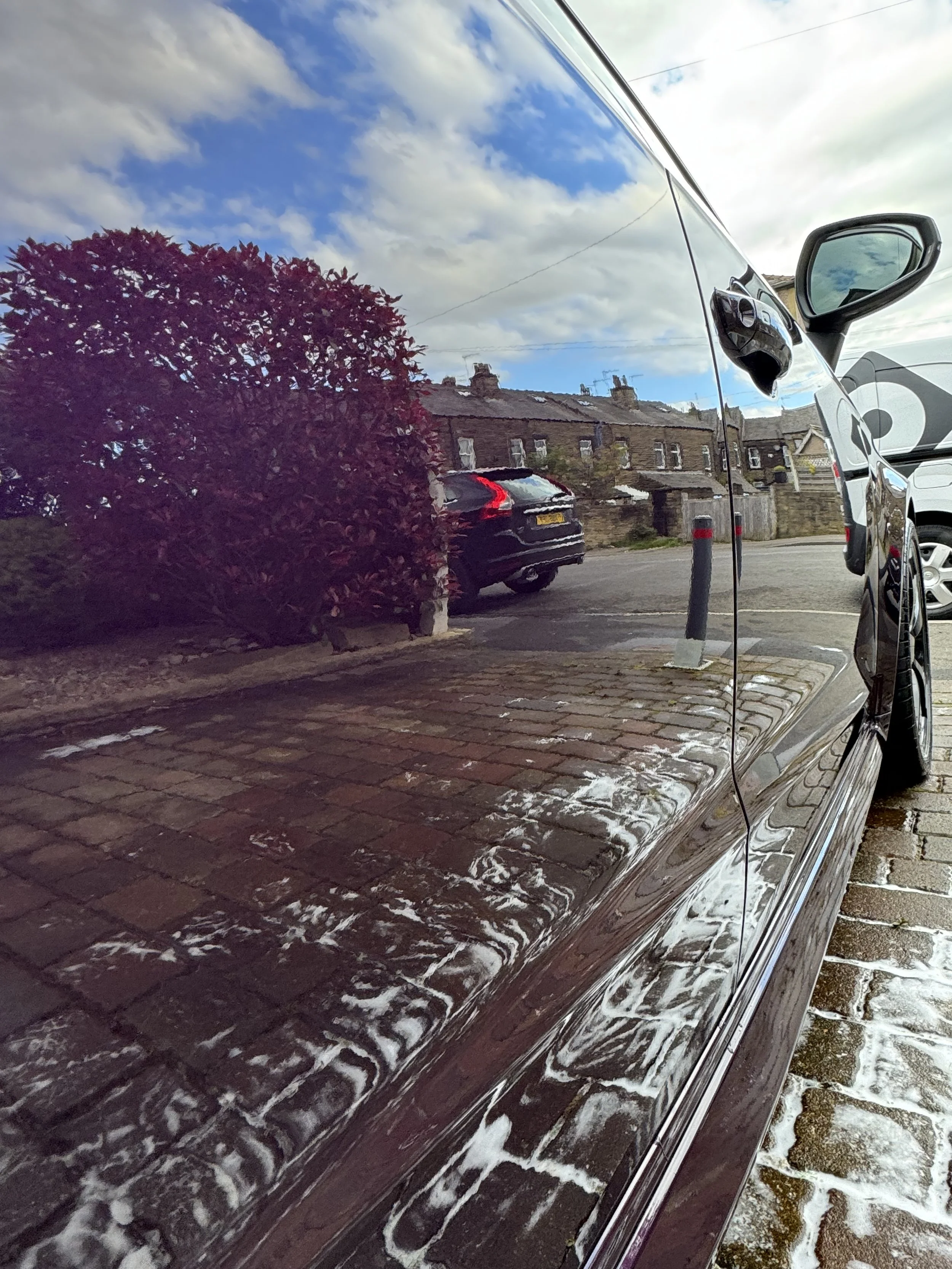 Reflection of houses, sky, clouds, and a tree on a black car parked on a brick-paved street, with soap suds visible on the car's surface.