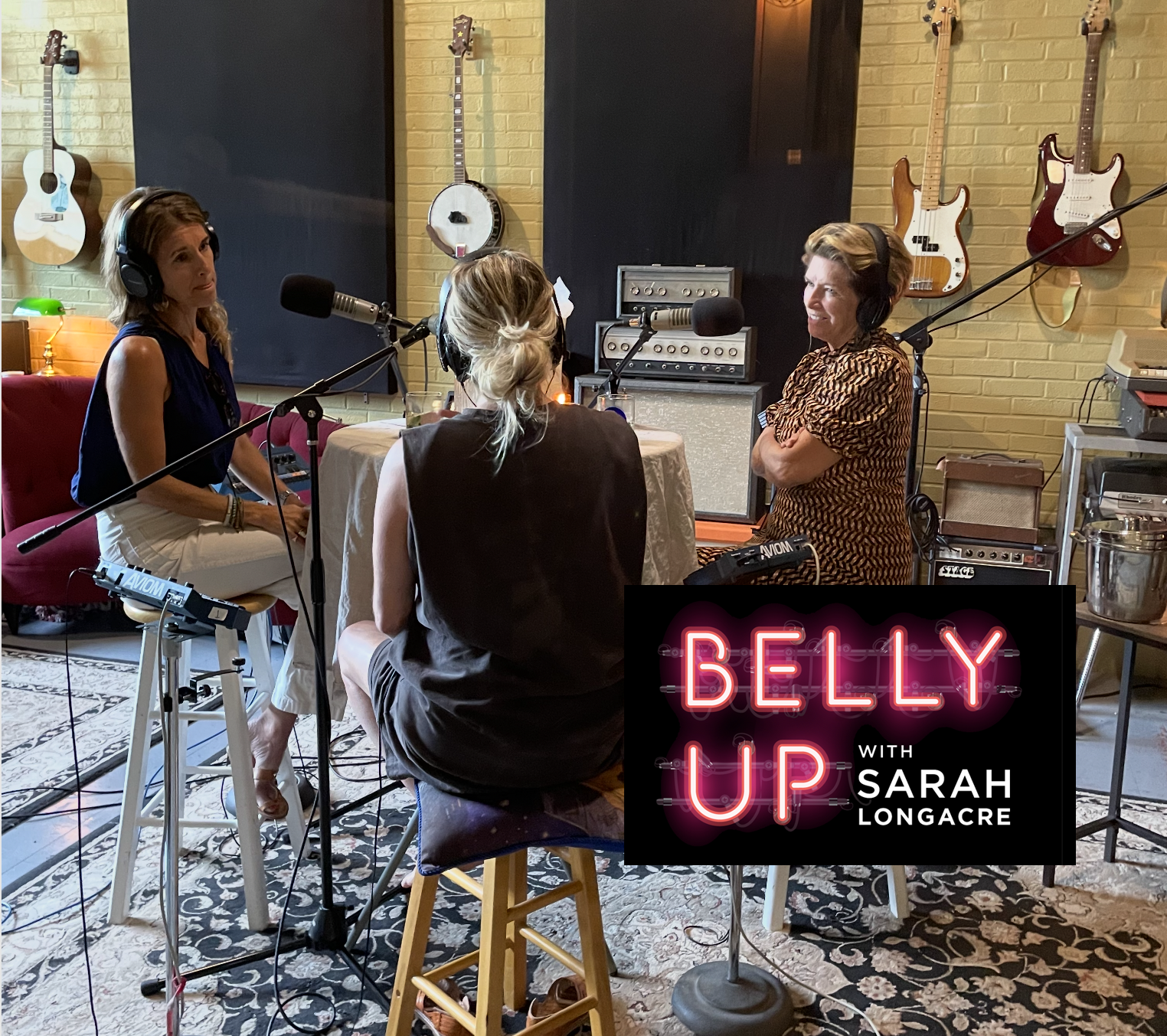 Three women participate in a podcast recording session in a studio with musical instruments on the wall and a sign reading 'Belly Up with Sarah Longacre'.