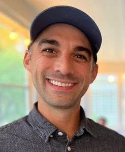 A smiling man wearing a blue baseball cap and a dark patterned shirt, standing indoors with a blurred background.