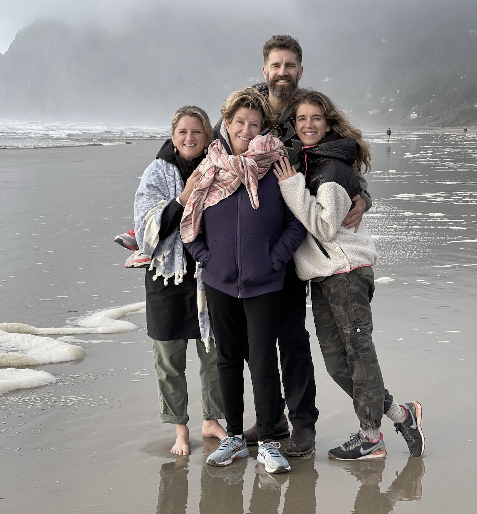 Group of five people hugging and smiling on a beach with foggy mountains in the background.