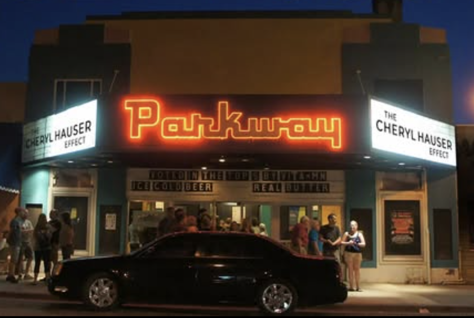Nighttime scene outside the Parkway theater showing a neon sign, a group of people standing outside, and a black car parked in front.