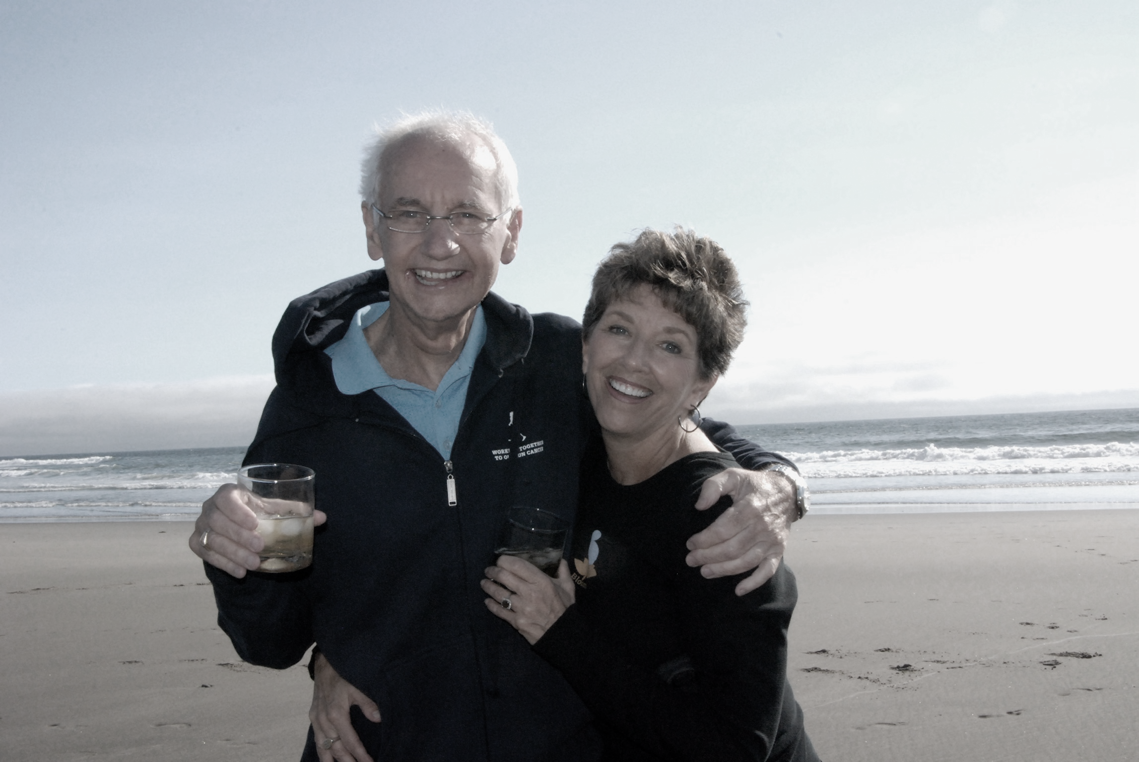 Older man and woman smiling on the beach, holding drinks, with ocean waves in the background.