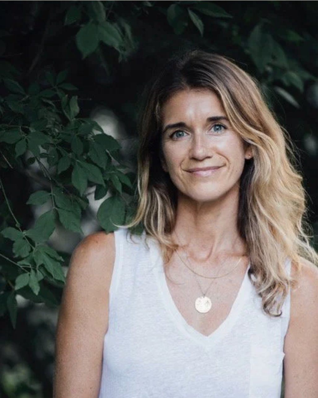 A woman with wavy blonde hair, blue eyes, and fair skin, smiling outdoors against green leafy background, wearing a white sleeveless top and a gold necklace.