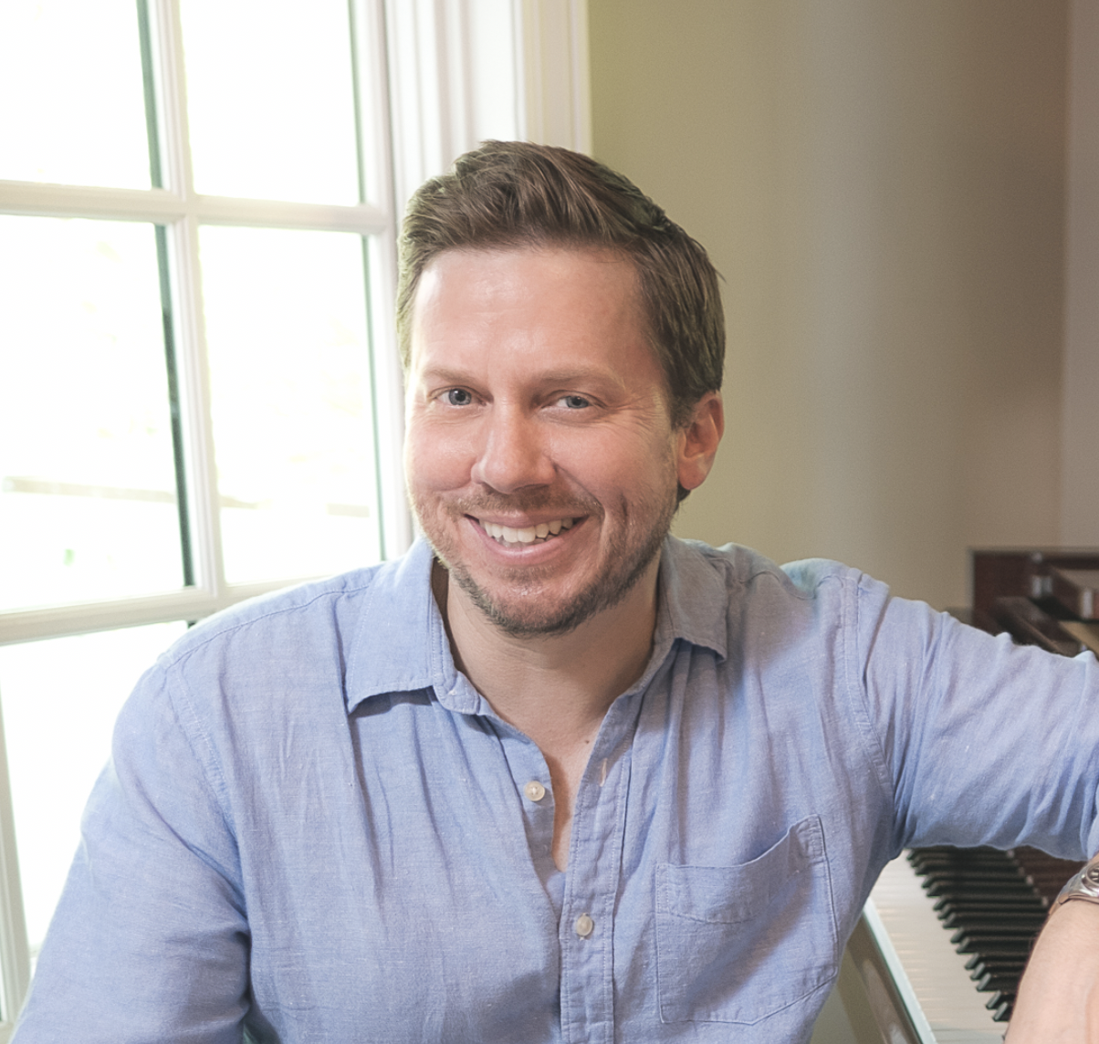 A smiling man with light brown hair, wearing a light blue shirt, sitting at a piano with a window in the background.