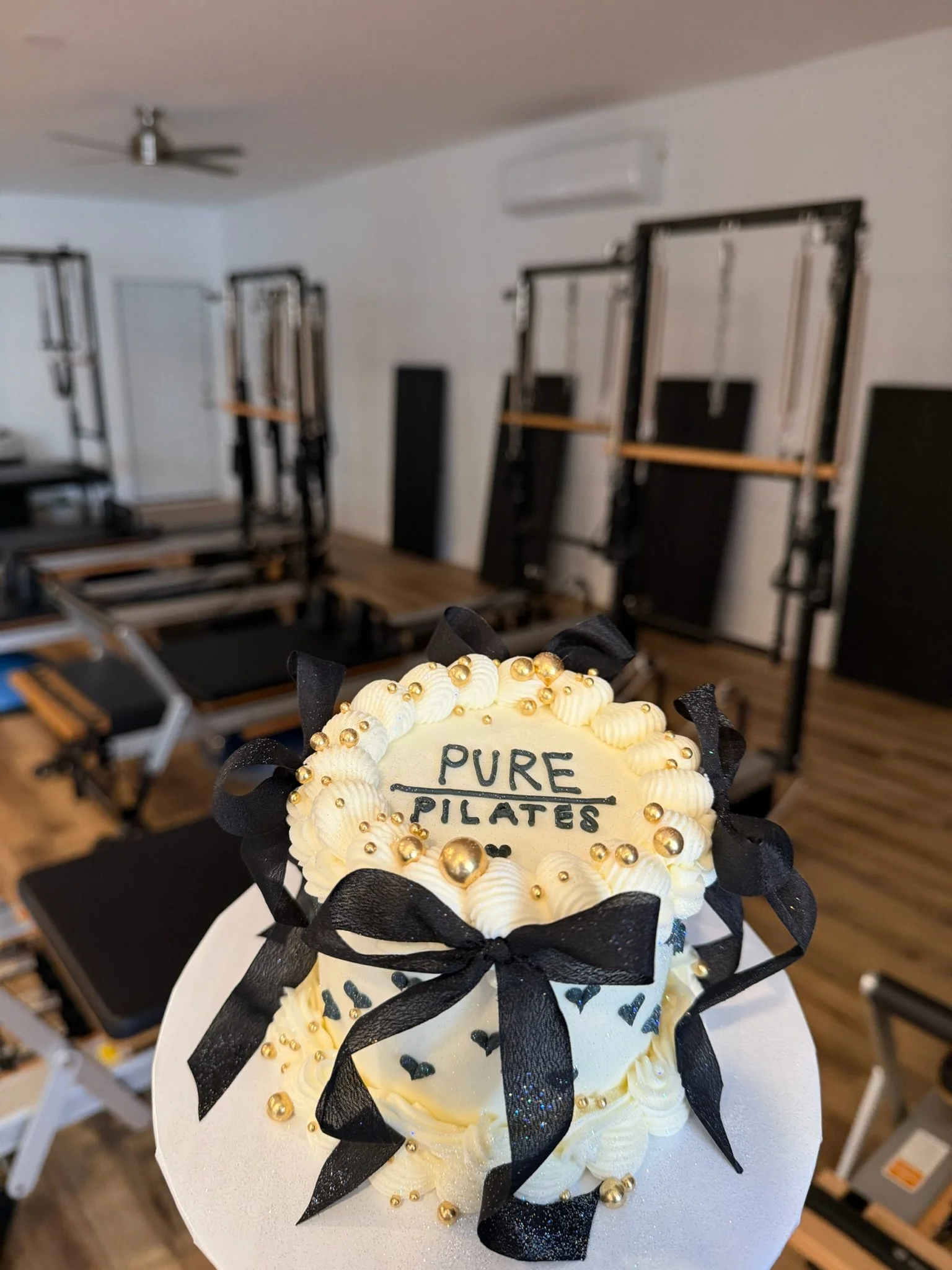 A cake decorated with white frosting, black ribbon, gold beads, black hearts, and a message that reads "Pure Pilates". In the background, there are Pilates reformer machines in a fitness studio with white walls and a ceiling fan.
