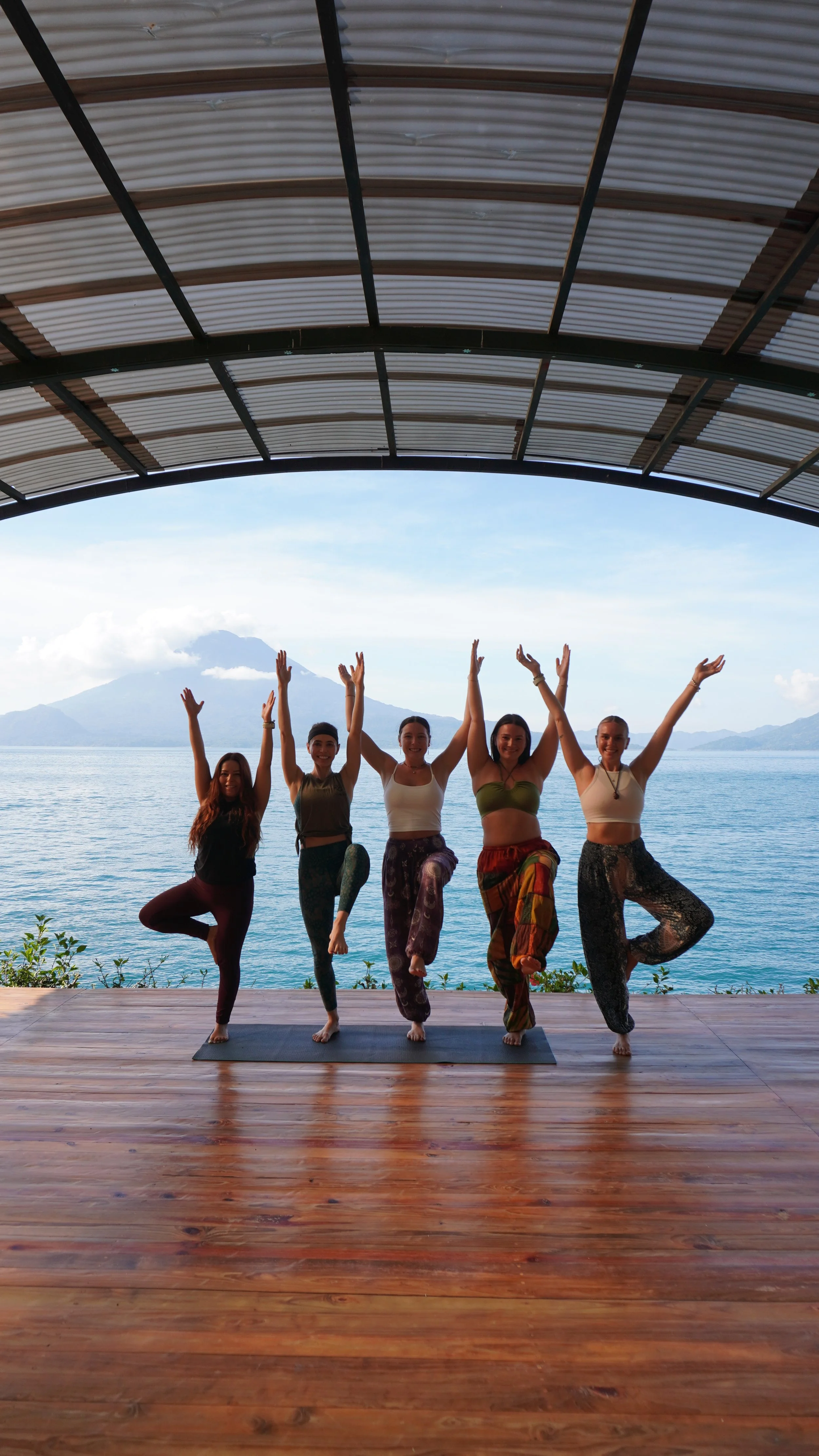 Five women practicing yoga outdoors near a body of water with mountains in the background, under a large metal and glass roof, standing on one leg with arms raised.