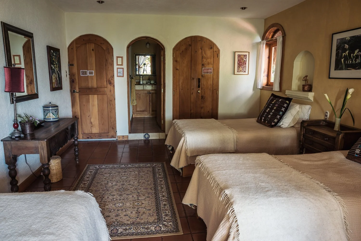 A cozy bedroom with two beds covered in beige bedspreads, wooden furniture, decorative wall art, and a small rug on a tiled floor. There are windows with curtains and an open door leading to a bathroom.