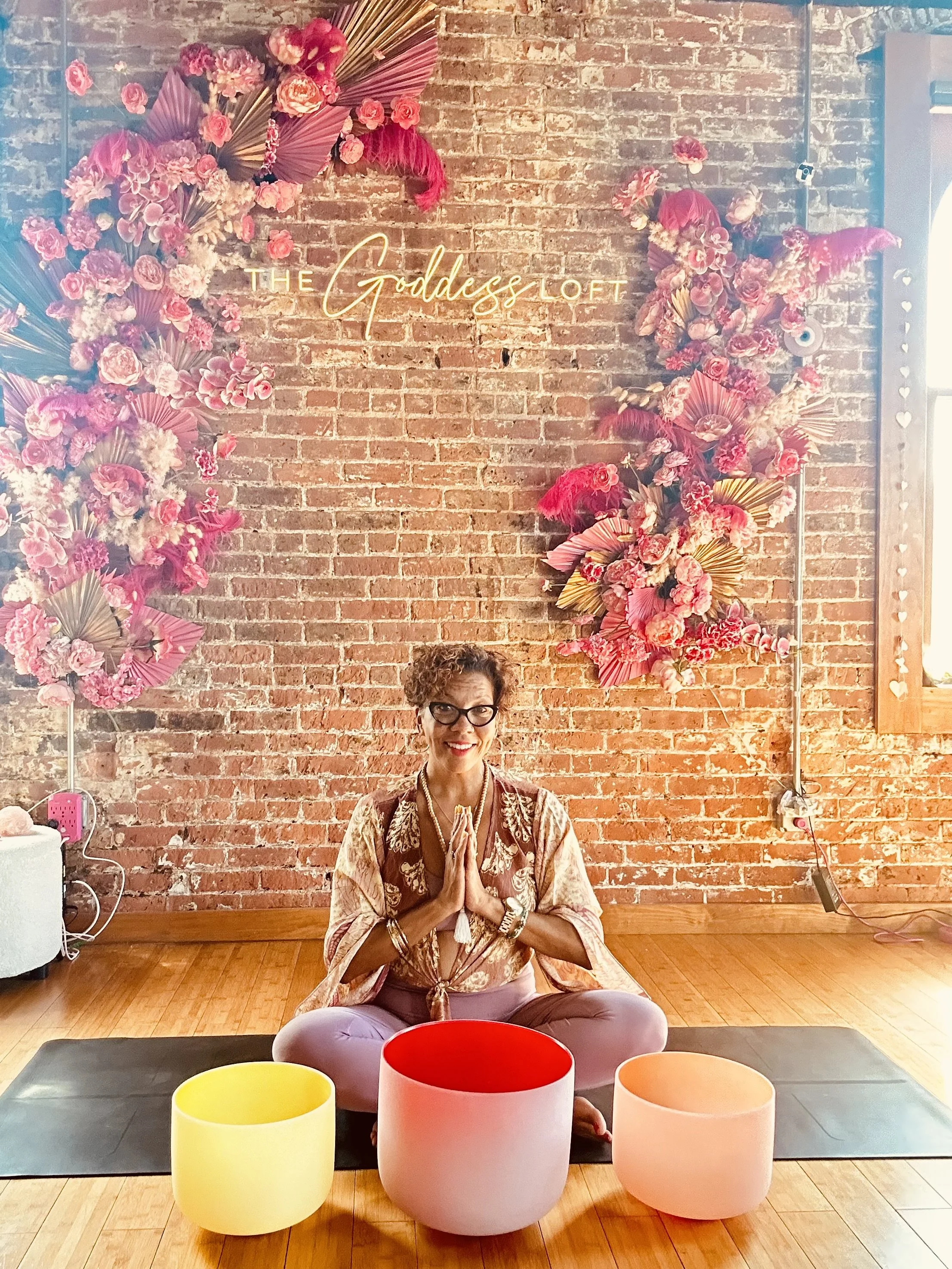 A woman practicing yoga or meditation in a seated cross-legged position on a yoga mat indoors against a brick wall decorated with pink and purple flowers and a neon sign that reads 'The Goddess Loft.' The woman has her hands pressed together in a prayer gesture, wearing glasses, a patterned top, and jewelry, with colorful large candles placed in front of her on the floor.
