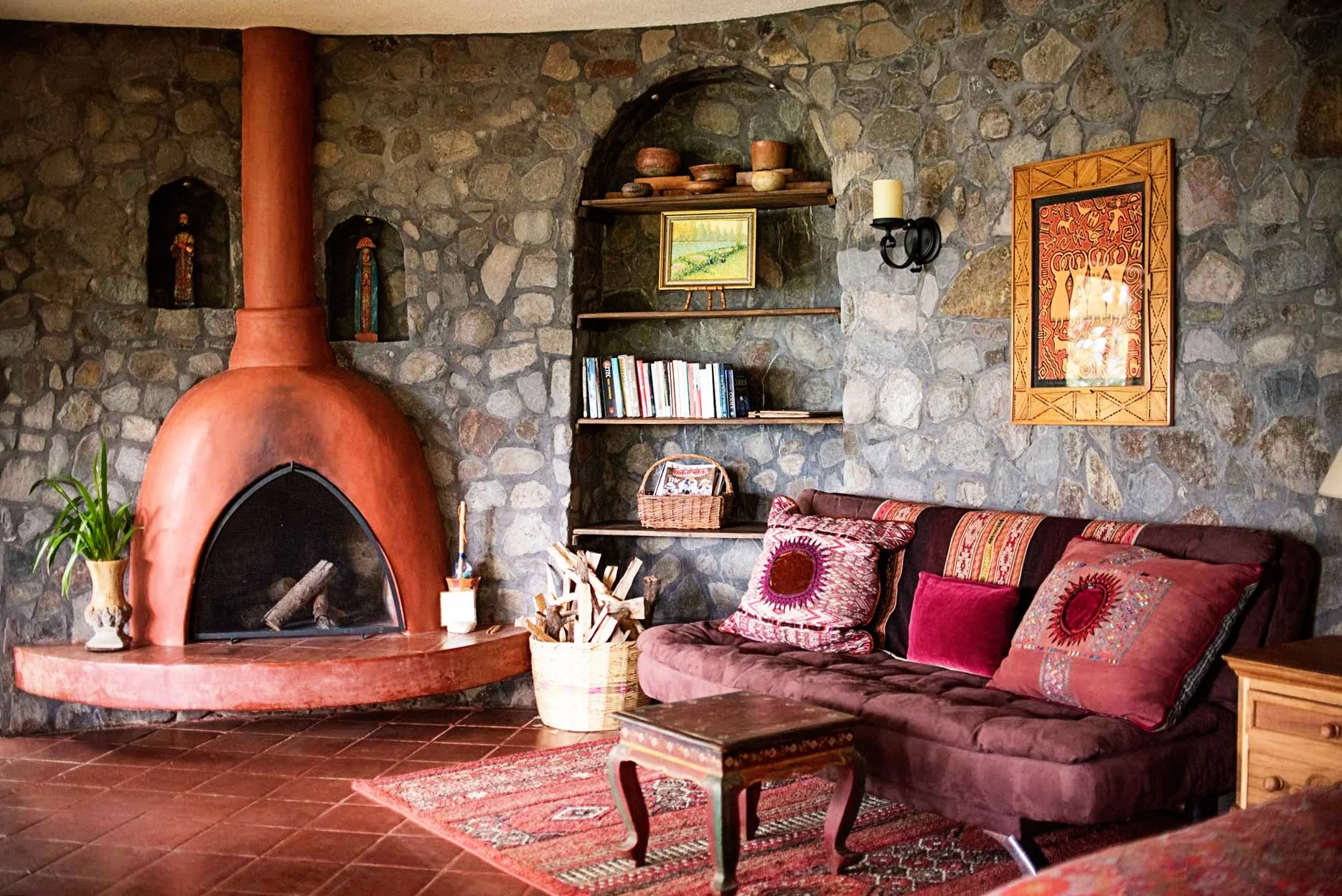 Cozy living room with a stone wall, a red adobe fireplace, bookshelves with books and pottery, a painting and tribal art, a wall sconce, a cushioned sofa with colorful patterned pillows, a small wooden table, a basket of firewood, and a potted plant.