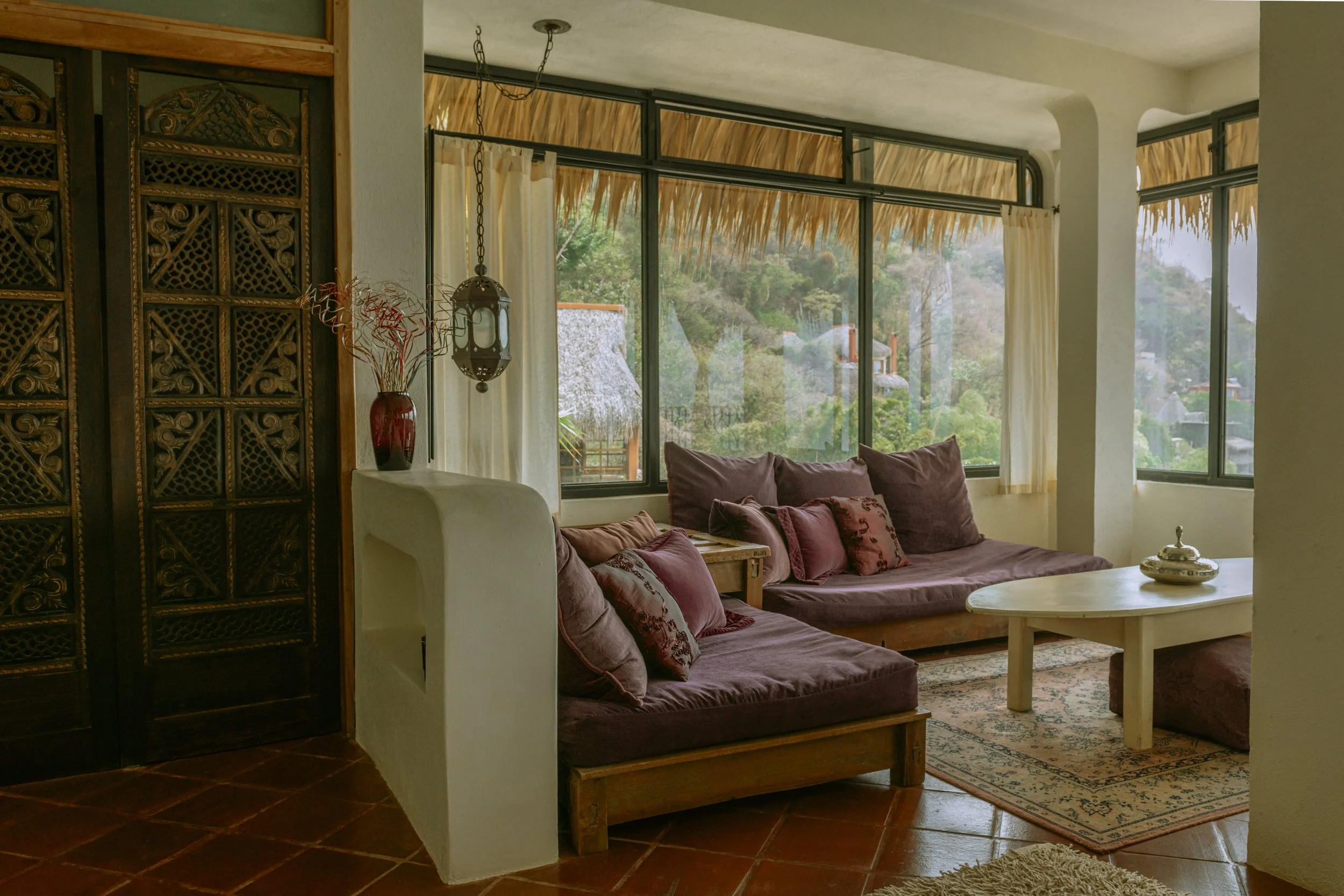 Living room with a cushioned sofa, a coffee table, large windows revealing a lush green landscape, and a decorative hanging lamp.