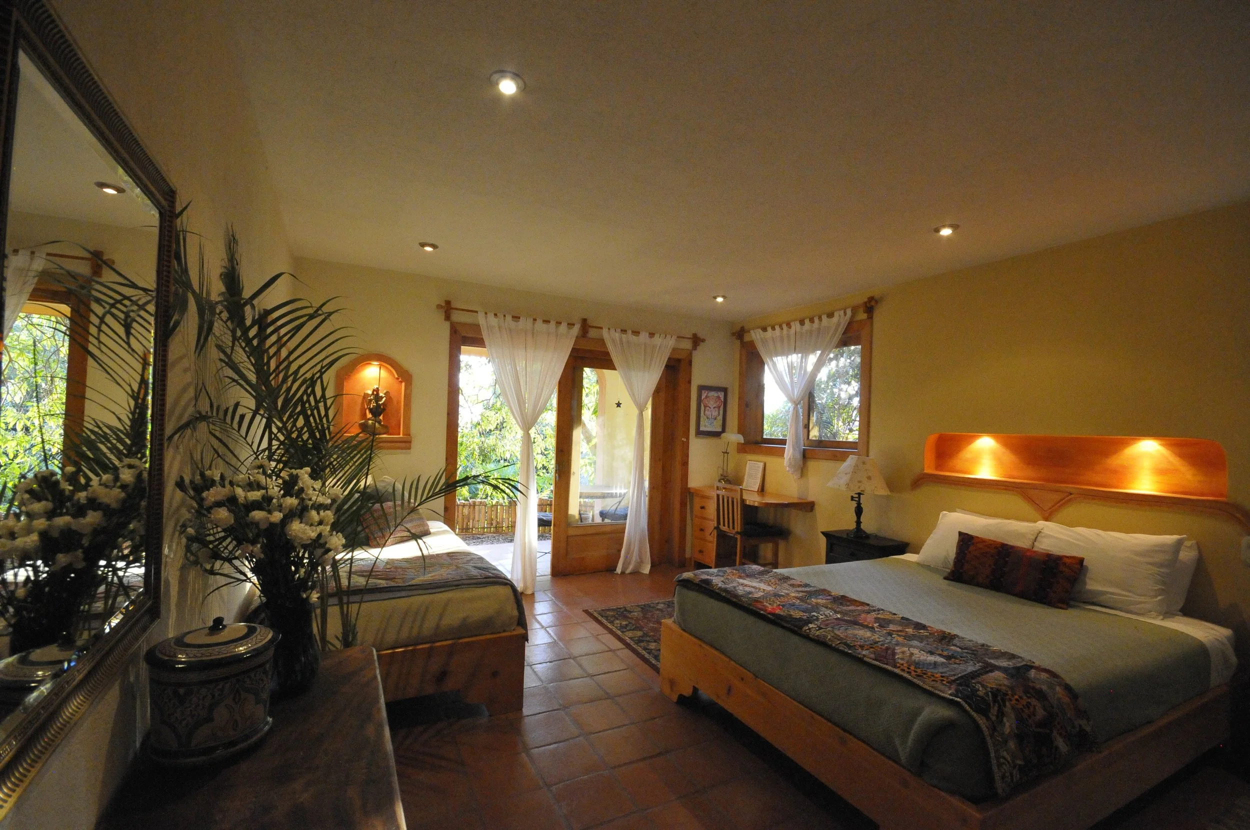 A cozy bedroom with two wooden beds, each with white linens and decorative pillows. There are large windows with sheer white curtains, allowing natural light to fill the room. A wooden desk, a lamp, and framed artwork adorn the walls. A mirror reflects a potted plant, and the room features warm wooden accents and tiled flooring.