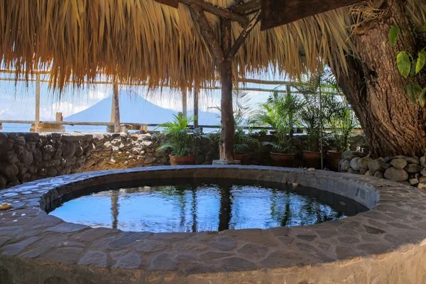 A stone hot tub or small pool under a thatched roof with tropical plants and mountain view in the background.