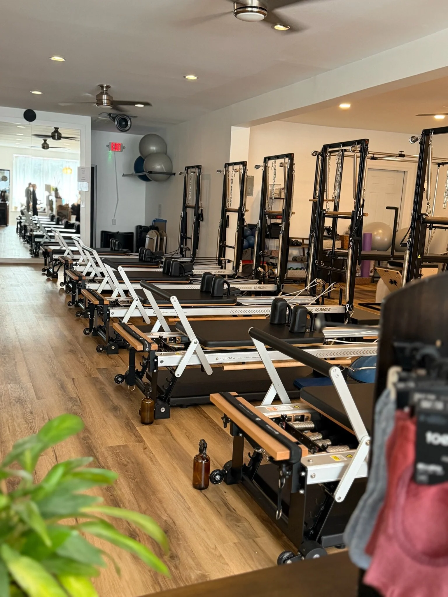 A gym room with pilates reformer machines arranged in a row on wooden flooring, exercise balls stored on shelves, and a mirror reflecting the room, with two people visible in the background.