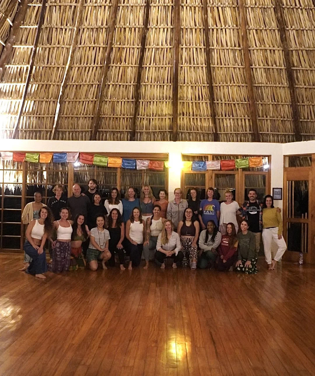 Group of people gathered inside a room with wooden floors and a thatched ceiling, decorated with colorful flags.