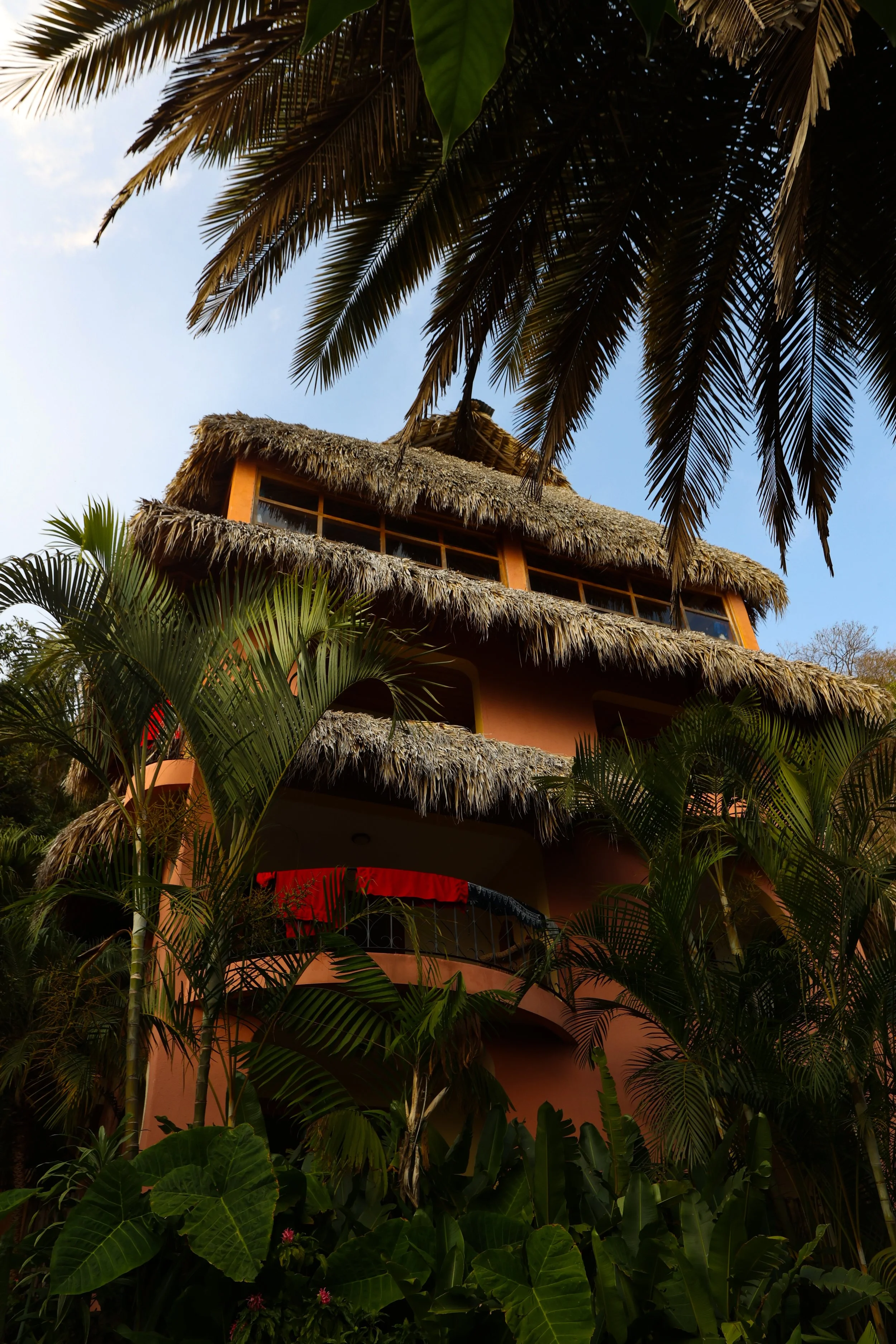 A multi-story building with a thatched roof, surrounded by lush tropical plants and trees, including palm trees, with a clear blue sky in the background.