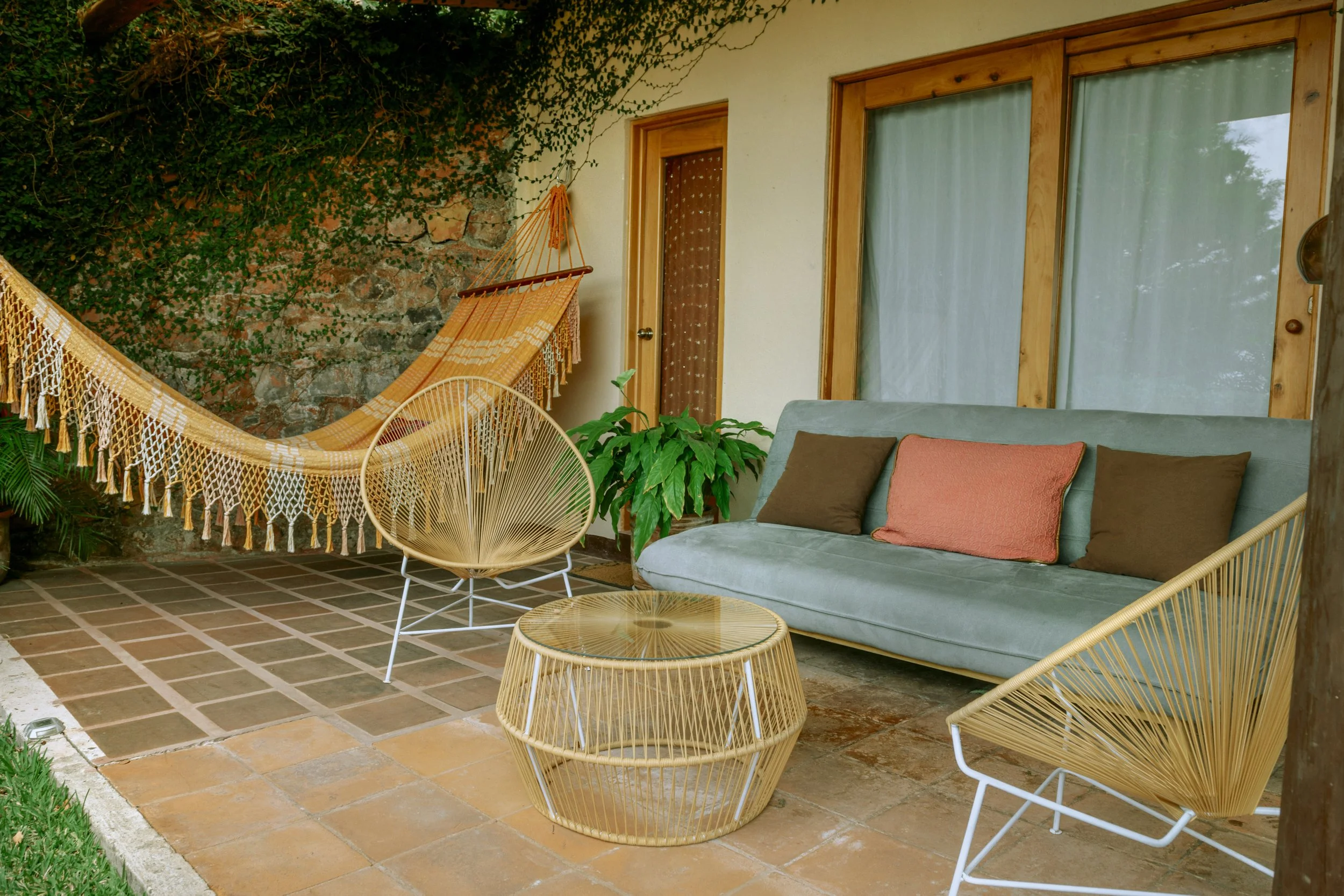 Outdoor patio with beige couch, orange and brown pillows, two wicker chairs, a small round table, a hammock, and green plants.