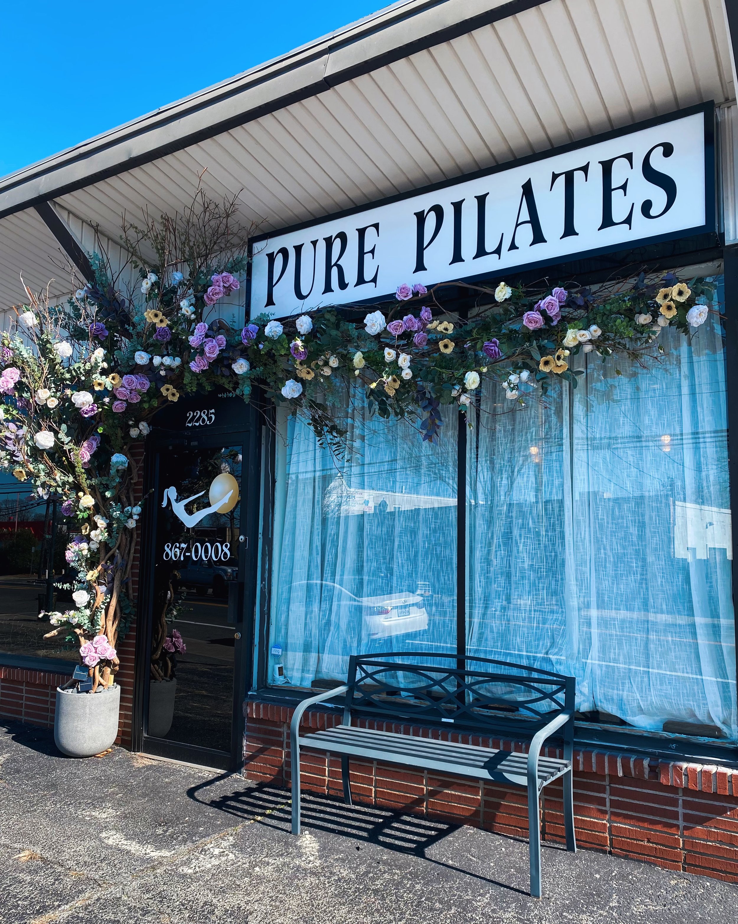 Storefront with a sign reading "Pure Pilates" above a glass door with a logo of a person doing a Pilates move. Decorative flowers surround the entrance, and a metal bench is in front of the window. The sidewalk and a bench are visible outside.