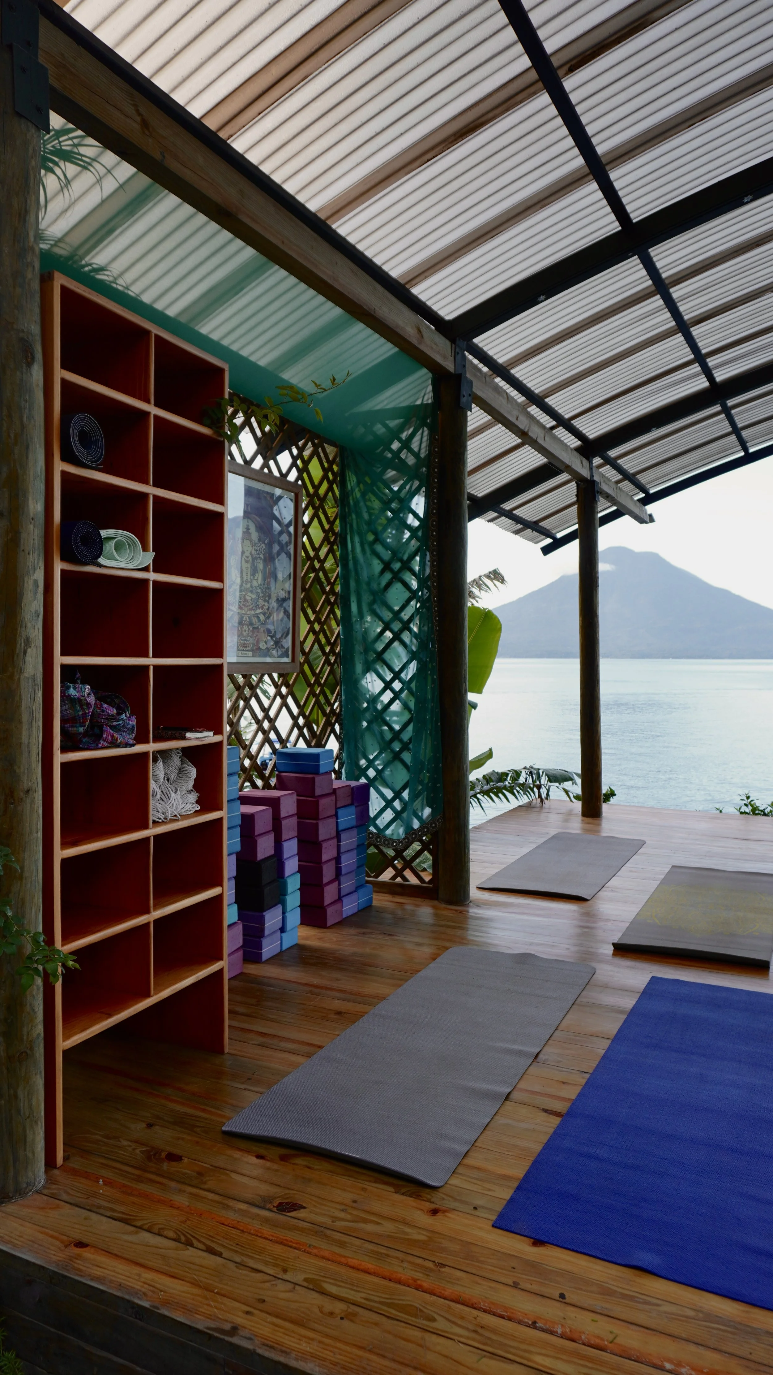 View of an outdoor yoga or meditation space on a wooden deck overlooking a body of water and mountains, with yoga mats arranged on the floor, a wooden shelf with rolled-up mats and other yoga equipment, and decorative elements like a framed picture and netting.