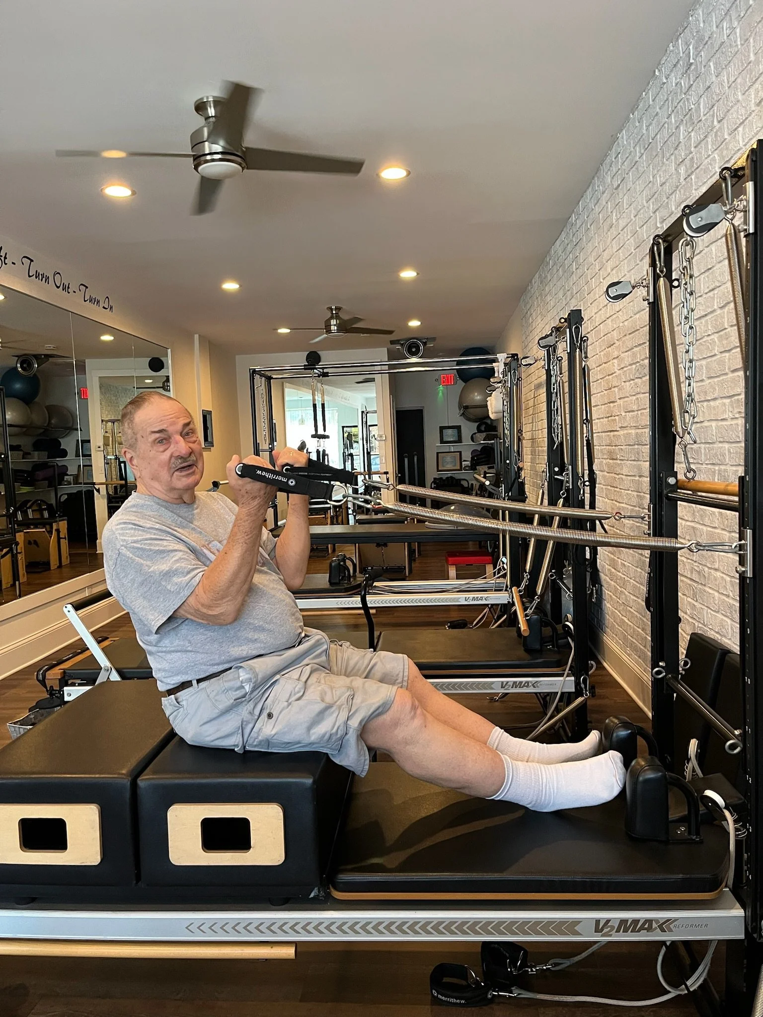 An elderly man with gray hair and mustache exercising on a Pilates reformer machine in a fitness studio, wearing a gray t-shirt and light-colored shorts, with the Pilates equipment and fitness equipment visible in the background.