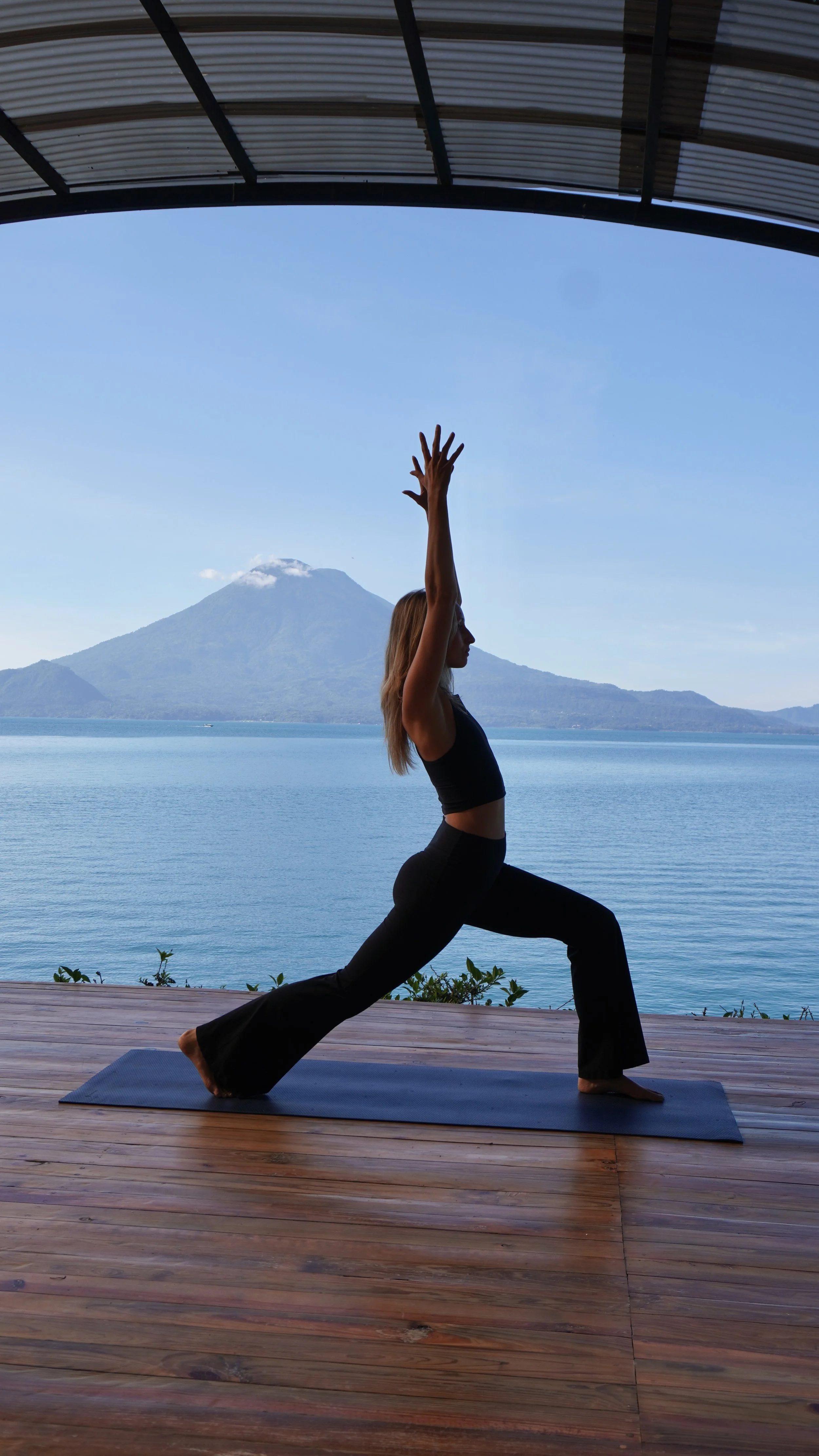 A woman practicing yoga outdoors on a wooden deck overlooking a lake, with mountains and a volcano in the background, wearing a black tank top and black pants.