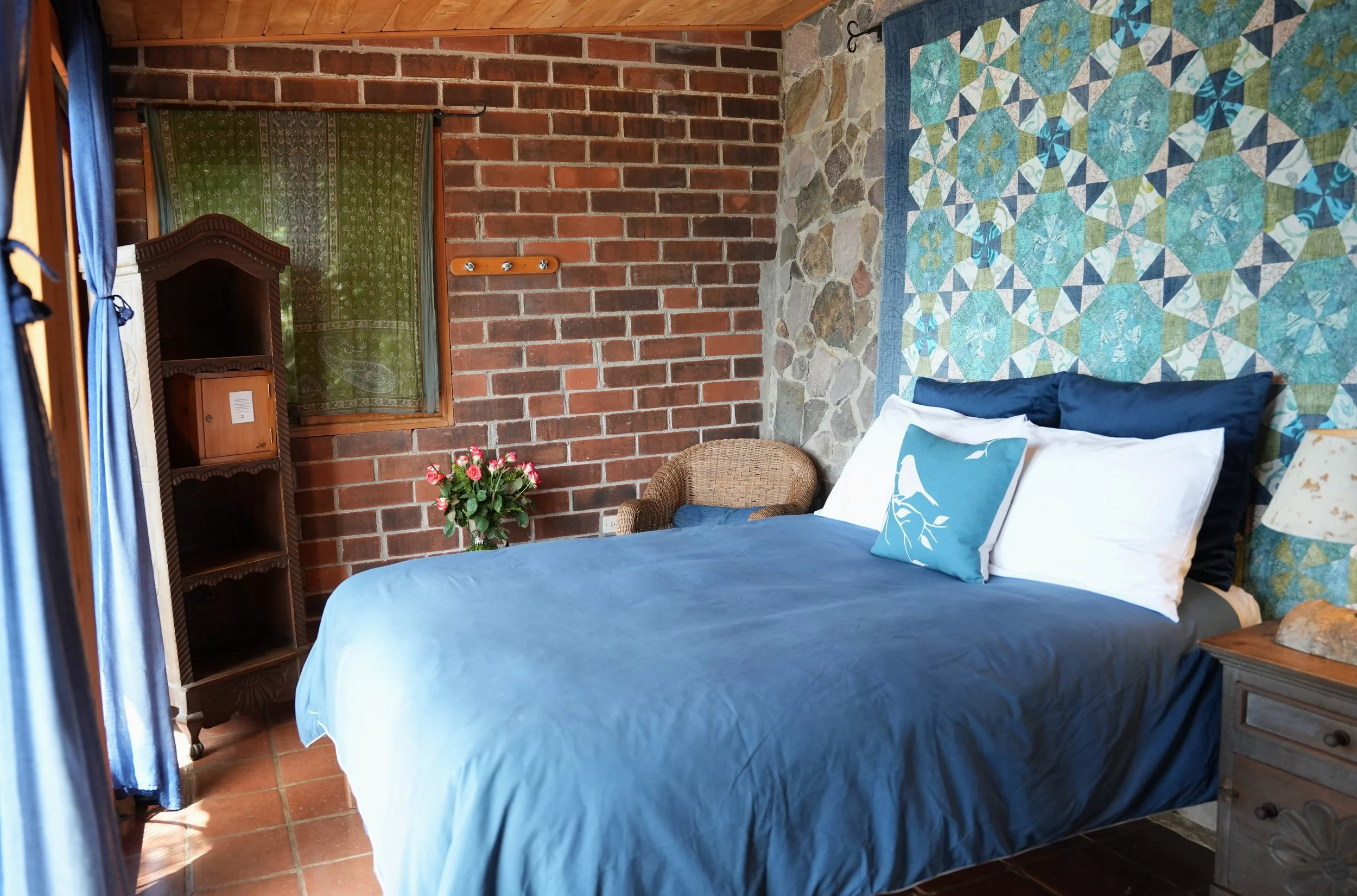 A cozy bedroom with a bed covered in a blue bedspread and decorative pillows, a brick accent wall, a window with green patterned curtains, a small wooden shelf, a wicker chair, and a bouquet of pink roses on a side table.