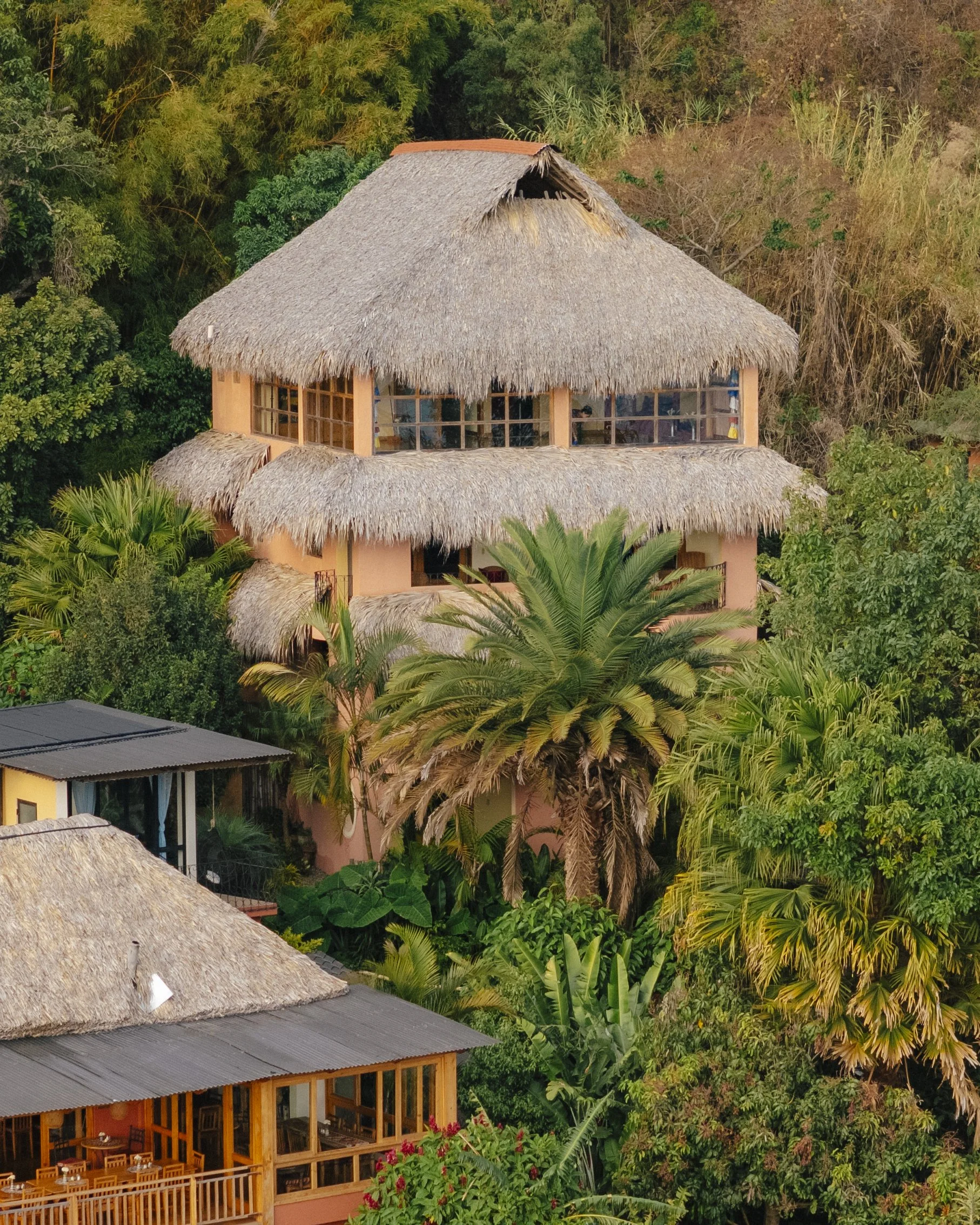 A multi-story tropical resort building with thatched roofs, surrounded by dense lush green trees and plants.