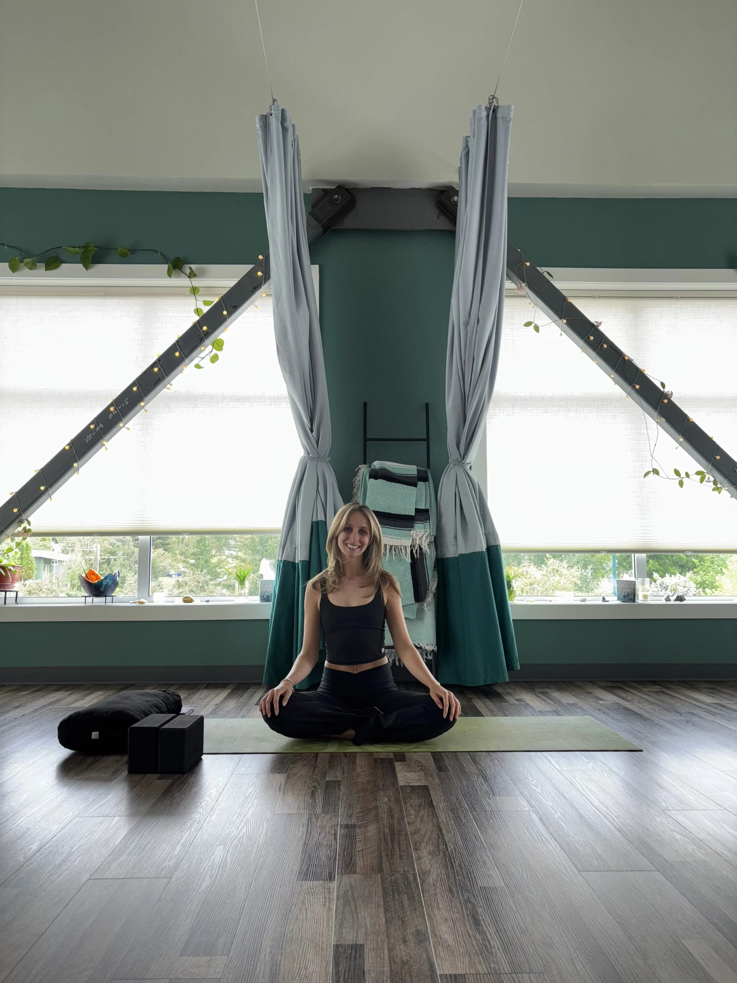 A woman practicing yoga in a living room with large windows, green walls, and a marquee uplit with fairy lights. She is sitting cross-legged on a yoga mat, smiling, with props and towels behind her and a gray curtain hanging from the ceiling.