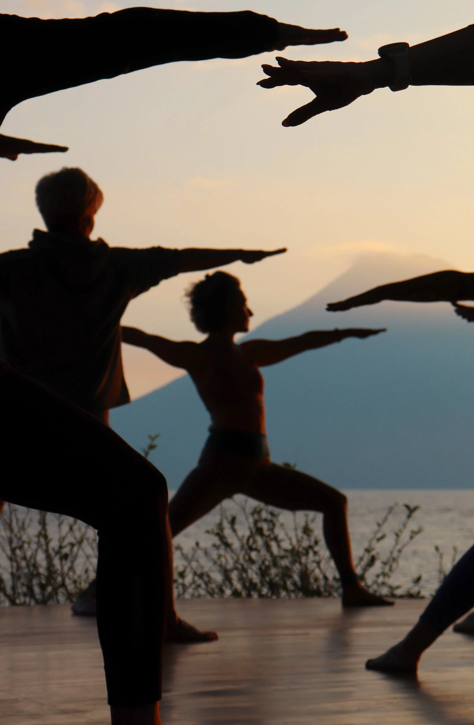 Silhouettes of children practicing yoga outdoors at sunset with mountains and water in the background.