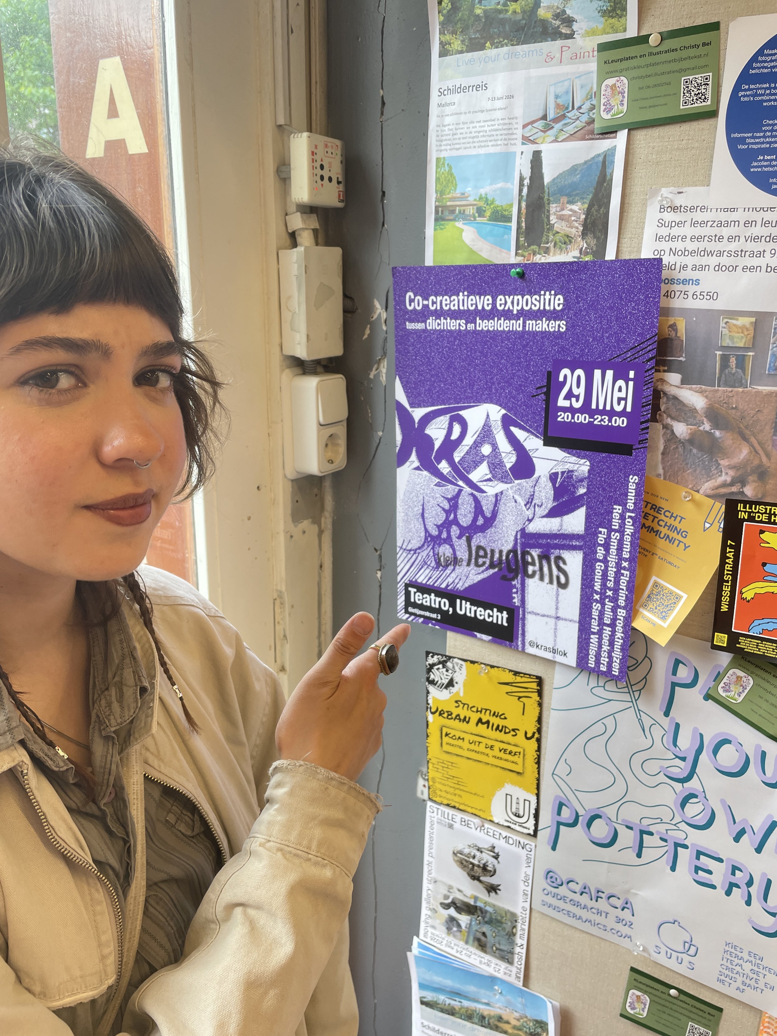 A woman with dark hair and a septum piercing pointing at a purple poster on a bulletin board with various other flyers and posters around her. The bulletin board has multiple colorful posters and flyers, some overlapping each other. The woman is wearing a beige jacket.