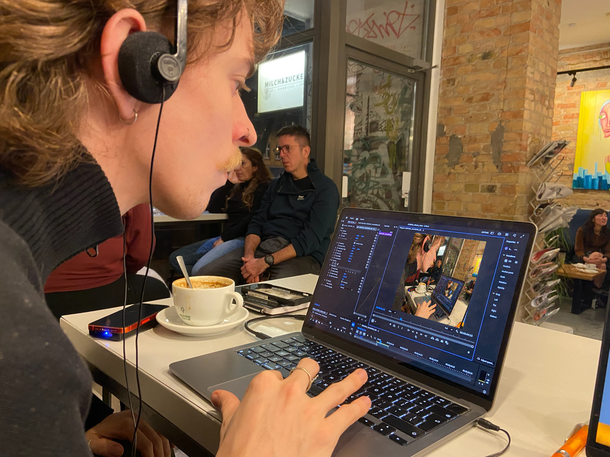 A person wearing headphones working on video editing software on a laptop in a coffee shop, with people sitting in the background and a colorful painting on the wall.