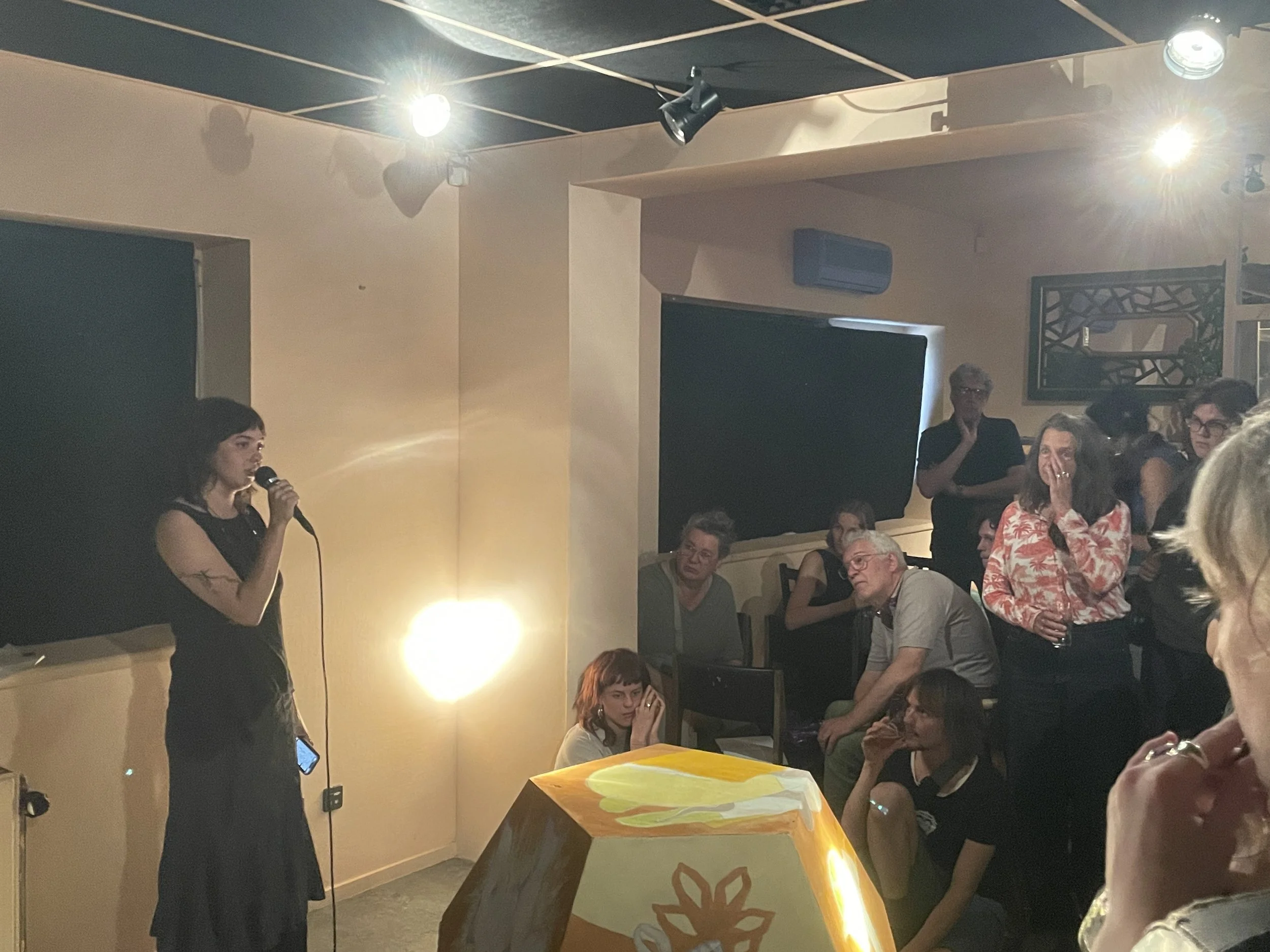A woman holding a microphone appears to be speaking or singing at a gathering. There are several people seated and standing around, some listening attentively. The setting looks like an indoor event or party with dim lighting and artificial ceiling lights.