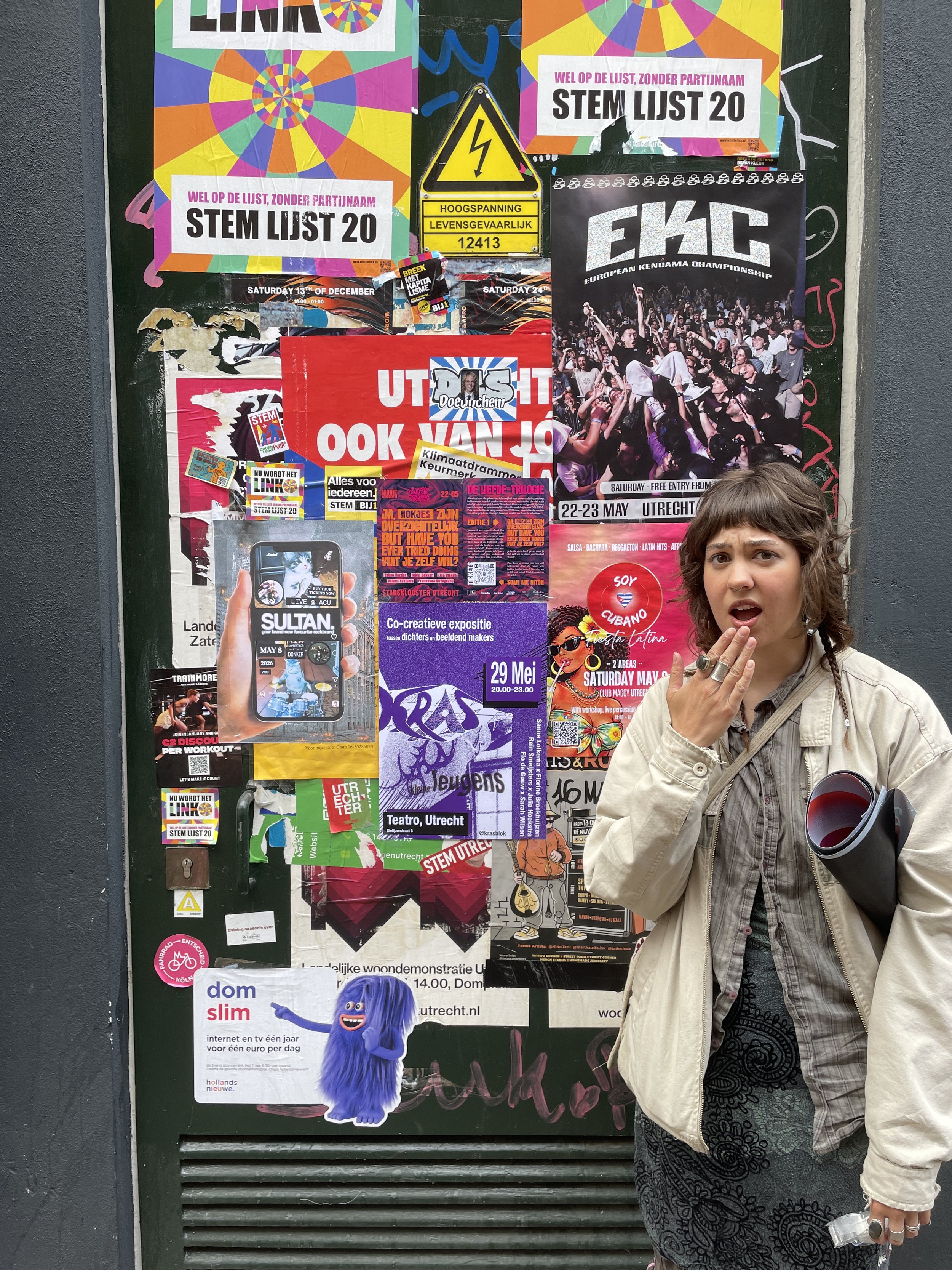 A large bulletin board covered with various colorful posters, flyers, and stickers, situated next to a gray wall. In front, a woman with short brown hair, wearing a beige jacket and black-patterned dress, is holding a black and red thermos and touching her face.