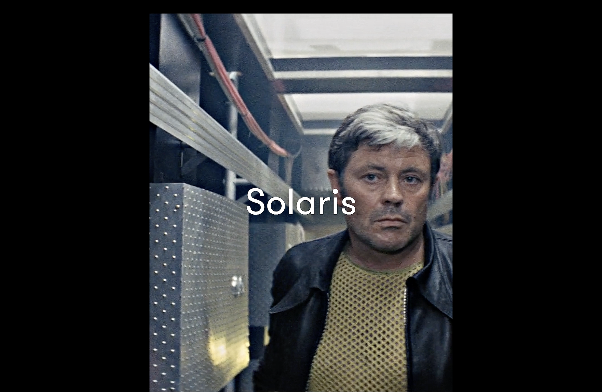 A man with gray hair and a serious expression, wearing a yellow mesh shirt and black leather jacket, standing in a dimly lit industrial or mechanical setting with metal equipment and pipes.