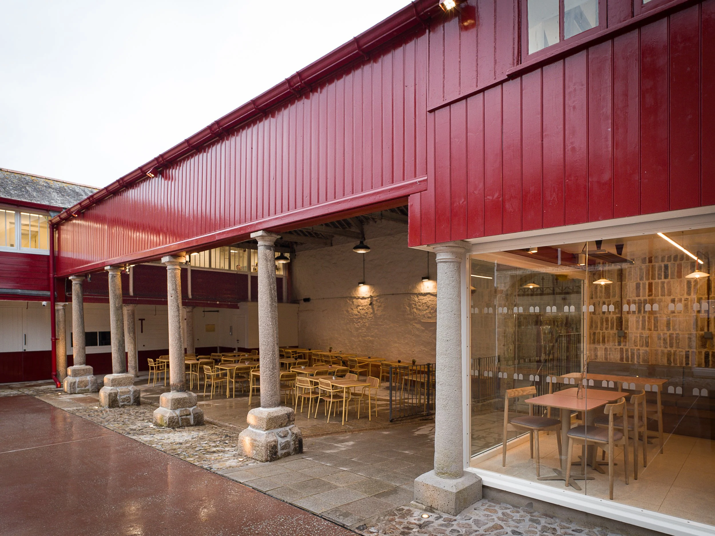 A red building with large glass windows and outdoor seating area with yellow chairs and tables underneath a covered porch, supported by stone columns, during dusk.