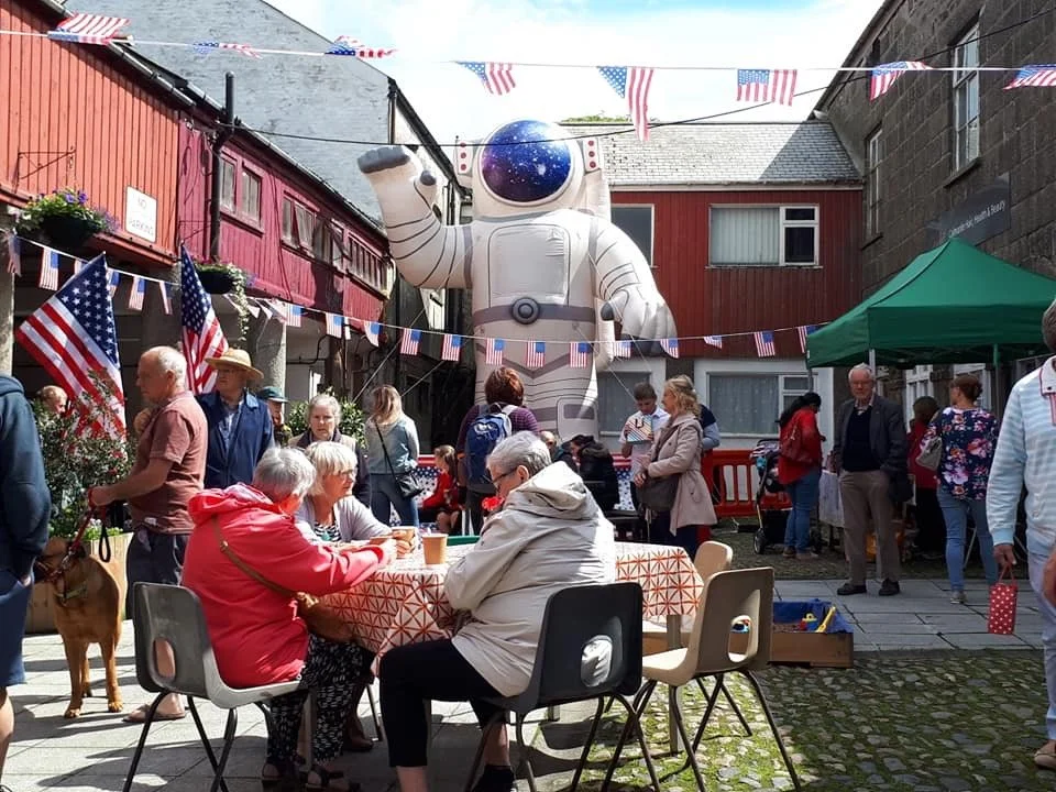 A street celebration with an oversized inflatable astronaut in the background, surrounded by people, American flags, and patriotic decorations.