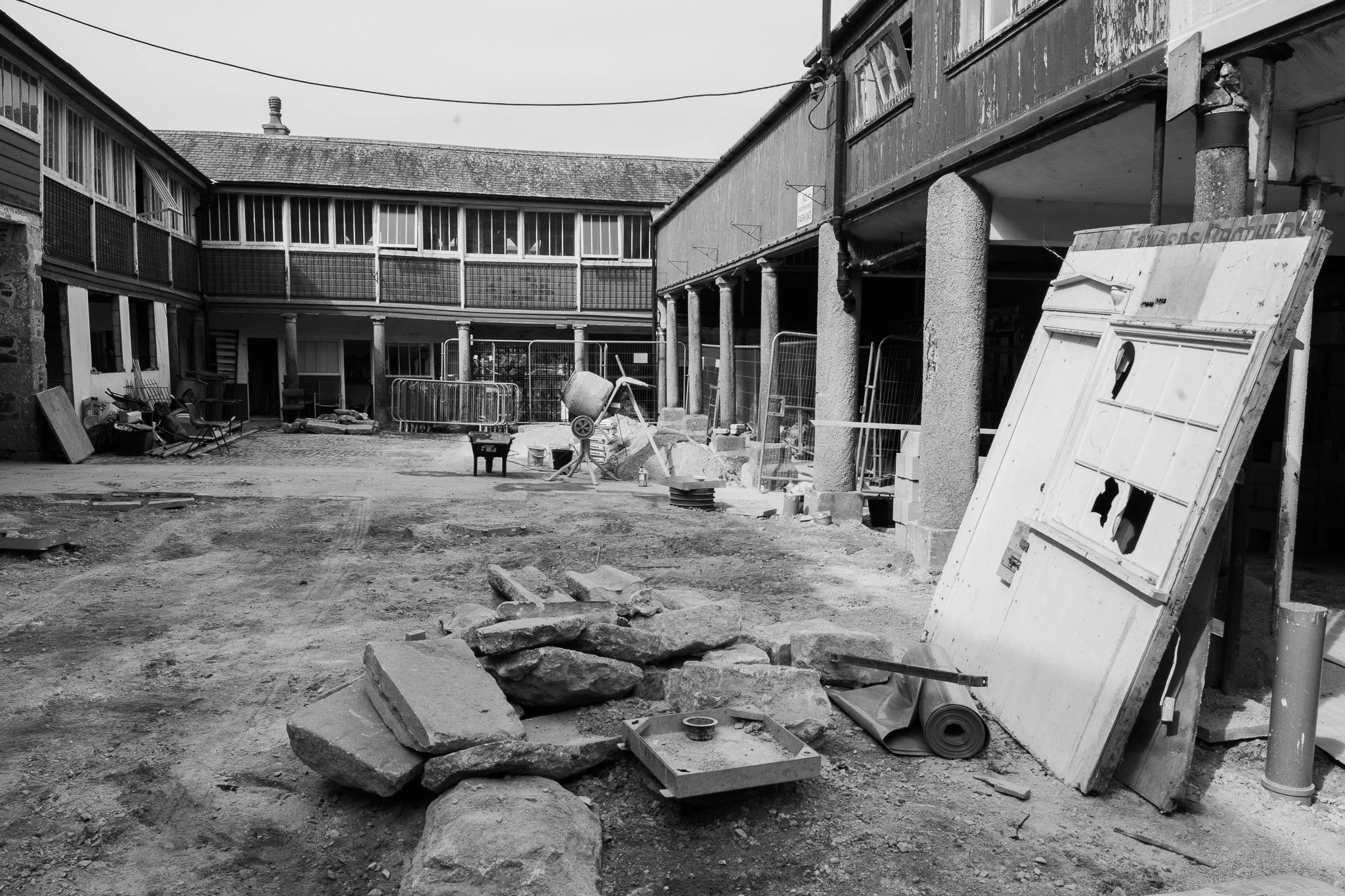 This is a black and white image of an outdoor construction or renovation site with scattered rocks, construction materials, and equipment. There are two levels of buildings with large windows, some of which are open, and a sign that says 'NO PARKING' is visible hanging from the upper level. Various construction supplies and tools are on the ground and leaning against structures. The area appears to be in disrepair or under construction.