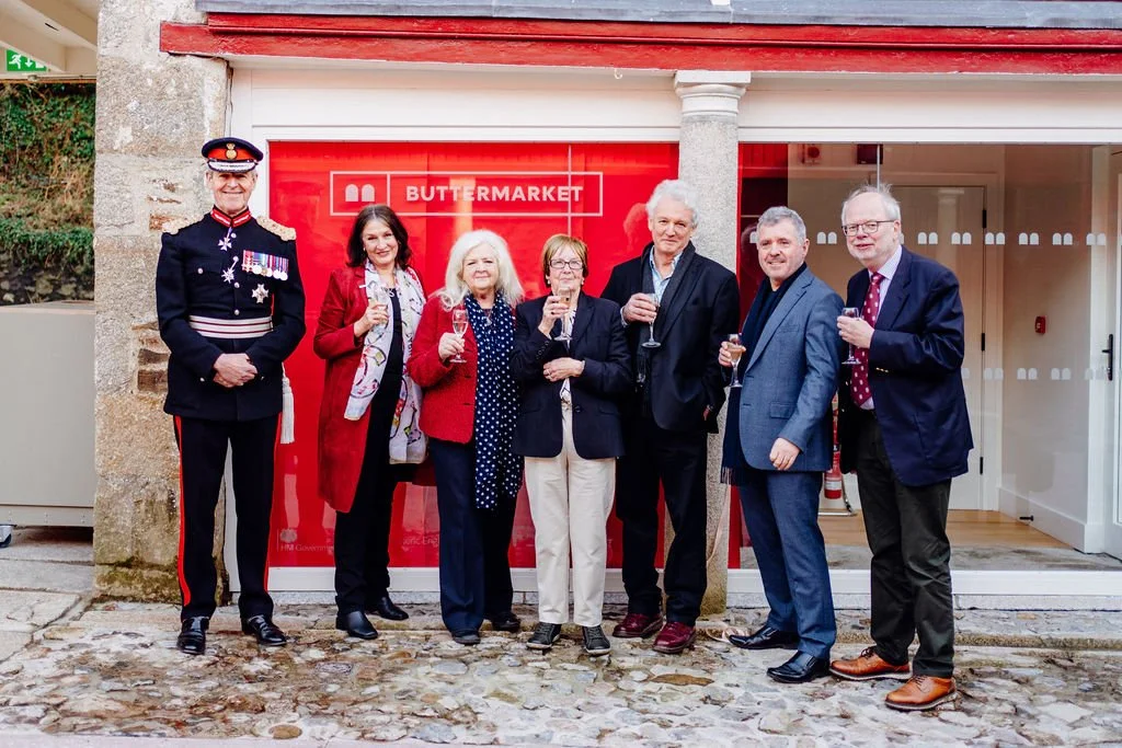 Group of seven people standing in front of a red store front labeled 'Buttermarket', holding champagne glasses and smiling. One person is dressed in a formal navy uniform with medals, while others are in business casual attire.