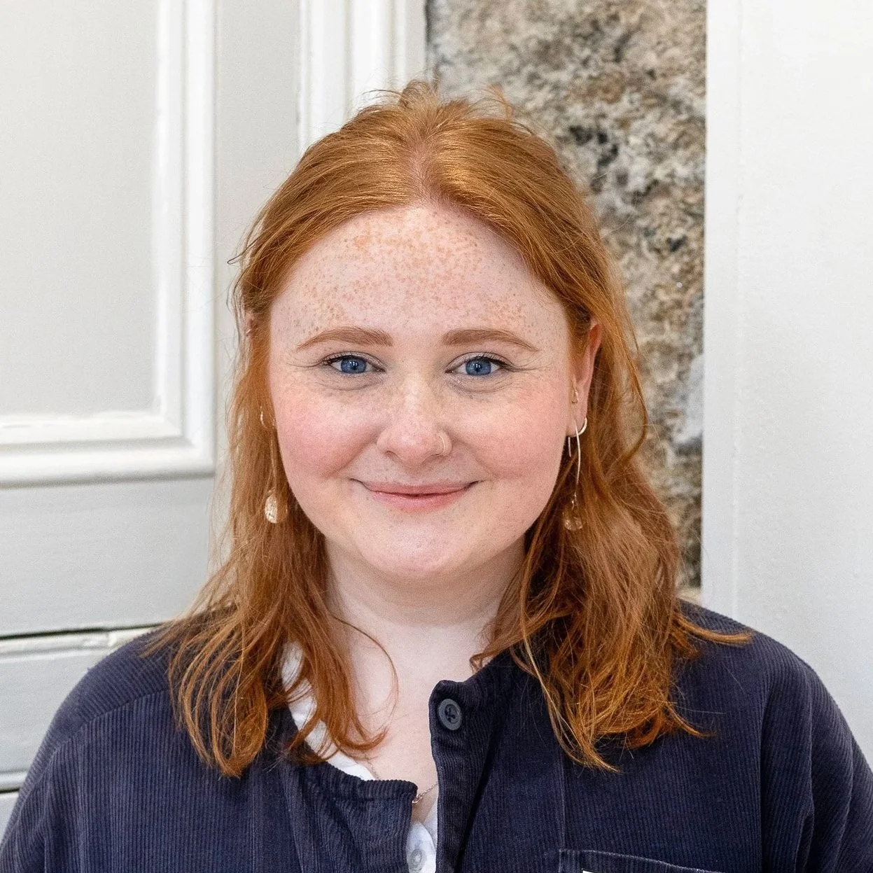 A woman with red hair and blue eyes smiling indoors, wearing a dark jacket and earrings.