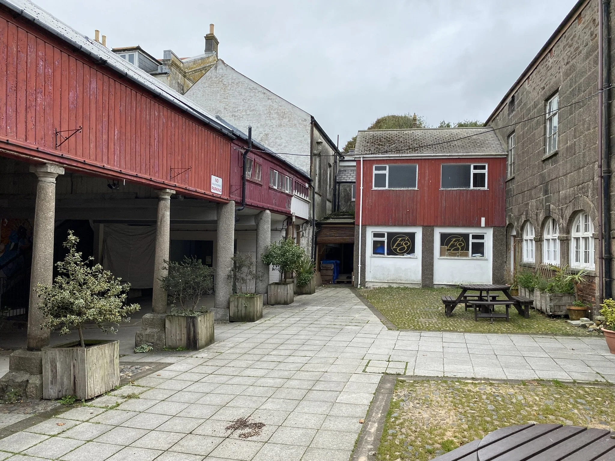 A courtyard with tiled and cobblestone paving, surrounded by old stone and red wooden buildings, some with windows and signs, small trees, benches, and plants.